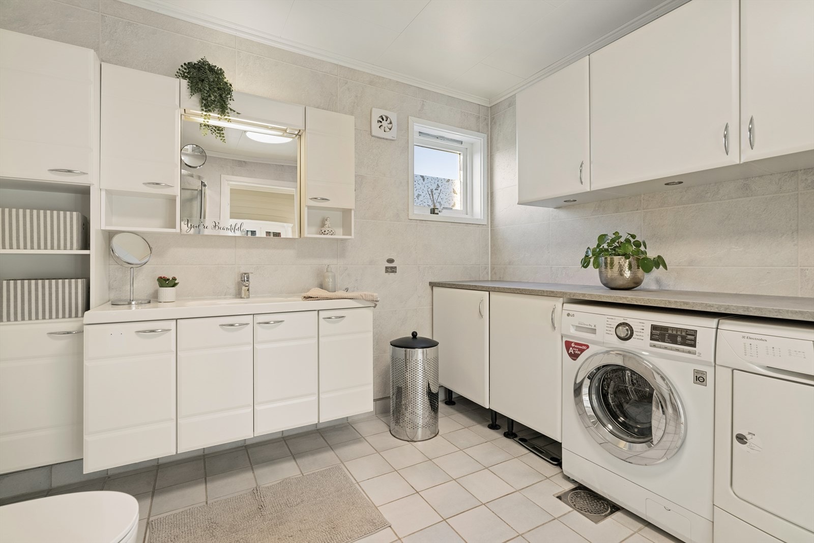 Innredningen er romslig med heldekkende servant, glatte fronter, speil med integrert belysning og tilhørende overskap. Det er montert en stor benkeplate i laminat over opplegg for vaskemaskin og tørketrommel. Galleribilde