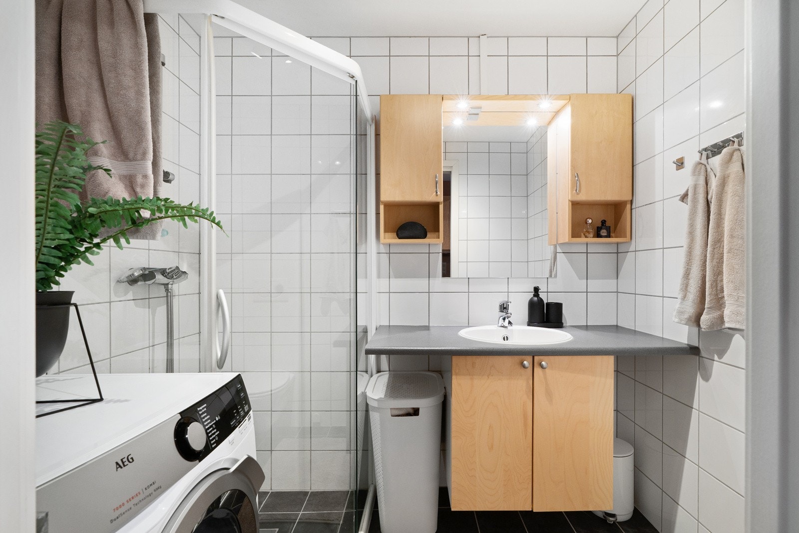 Baderommet er målt til 3,4 kvm, pent innredet med mørke, kvadratiske fliser på gulv og tilsvarende lyse fliser på vegg. Innredningen har lagringsplass i servantskap, supplert med veggmonterte skap, med integrert belysning. Galleribilde
