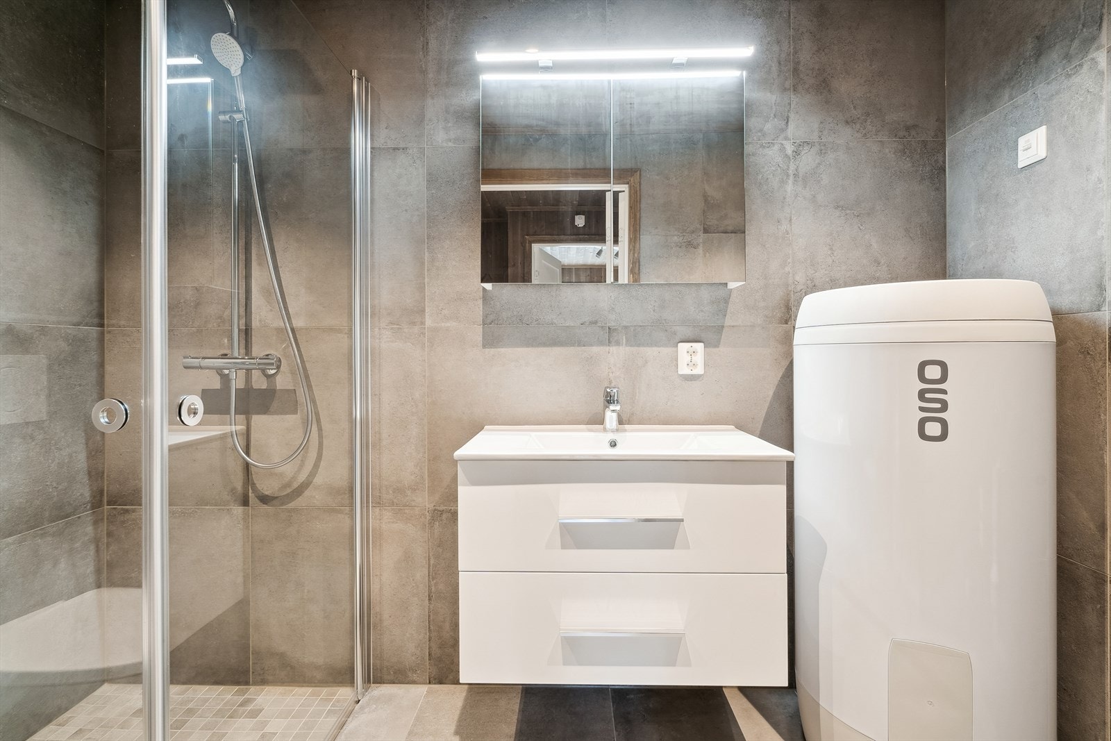 Badet inneholder dusjhjørnet med glassdører, servant med innredning, praktisk speilskap, vegghengt toalett og opplegg til vaskemaskin. Galleribilde