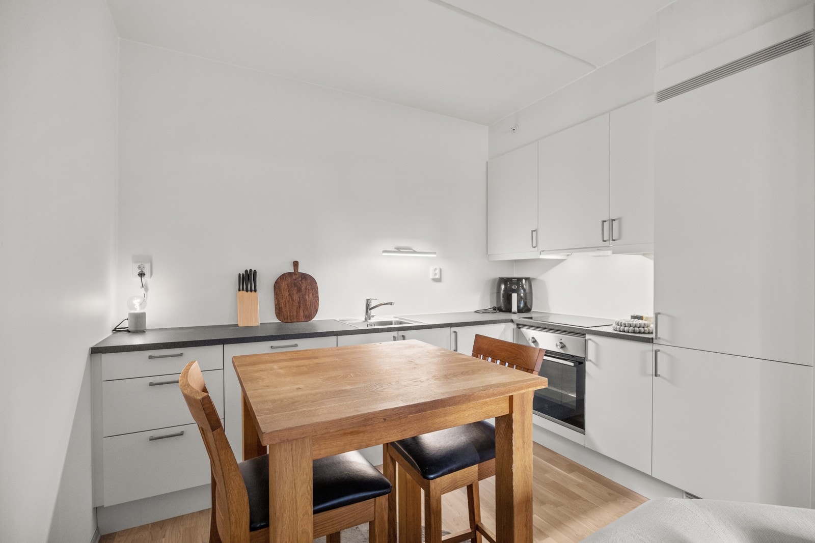 Pent kjøkken fra byggeår i L-form. Hvite underskap og overskap, samt belysning under høyskapene. Galleribilde