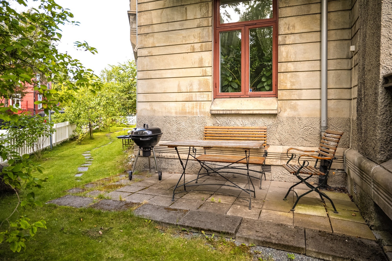 Felles uteplass med grill, bord og stoler på steinlagt område, som vender ut mot gaten, omgitt av gressplen og trær. Galleribilde