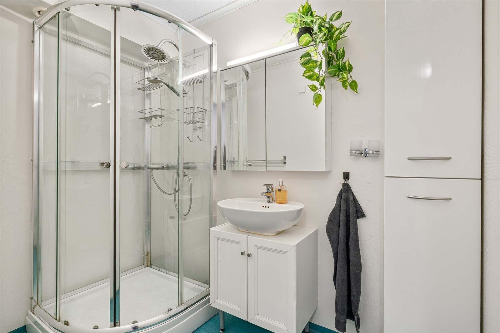 Lyst bad med dusjkabinett, servant og praktisk baderomsinnredning. Her får du en funksjonell løsning med god plass til oppbevaring, samtidig som de lyse flatene gir rommet et moderne og friskt preg - perfekt for en behagelig start på dagen. Galleribilde