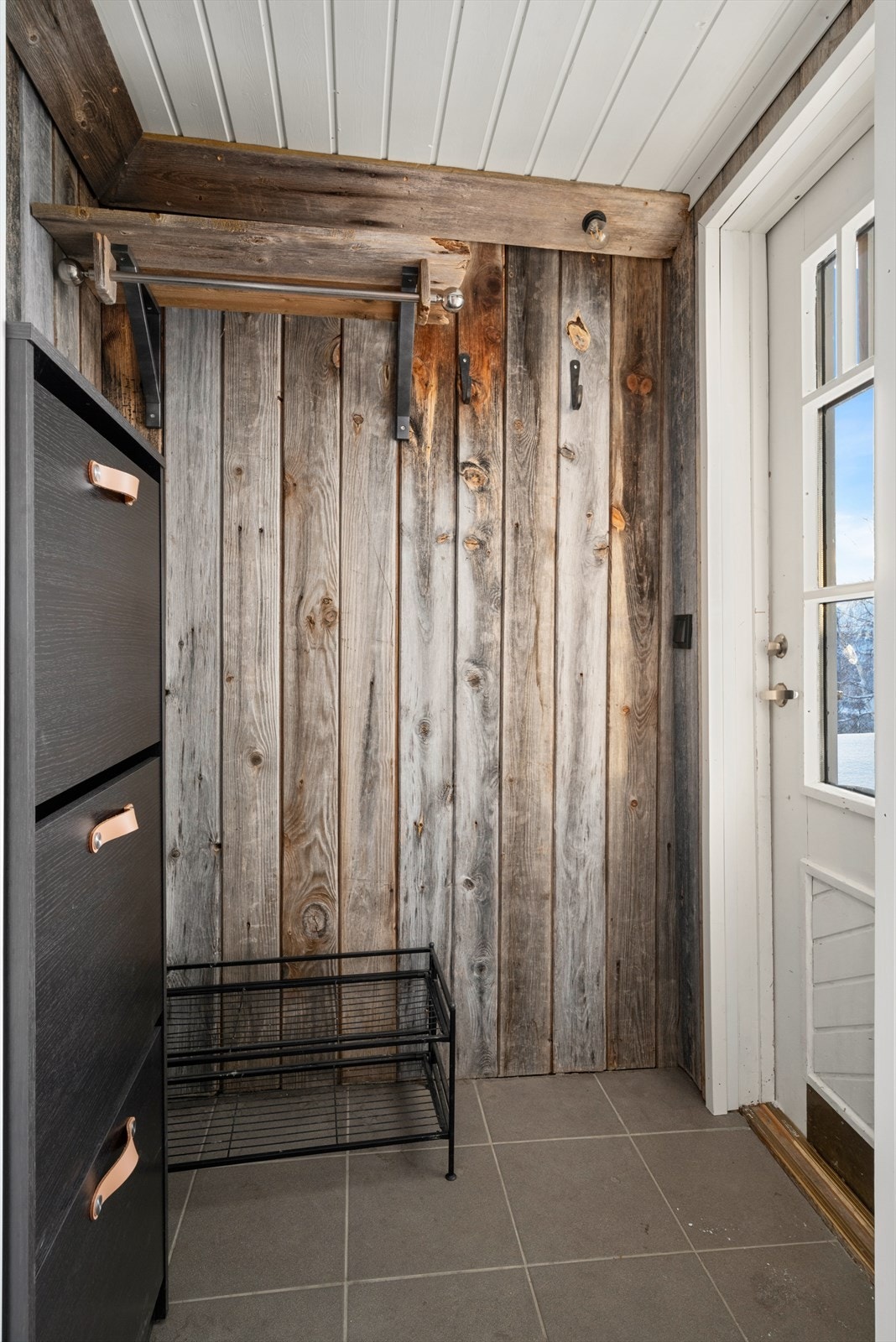 Vi blir ønsket velkommen i et vindfang på ca. 1,5 kvm med rustikk panel på veggene og grå fliser på gulvet. Galleribilde