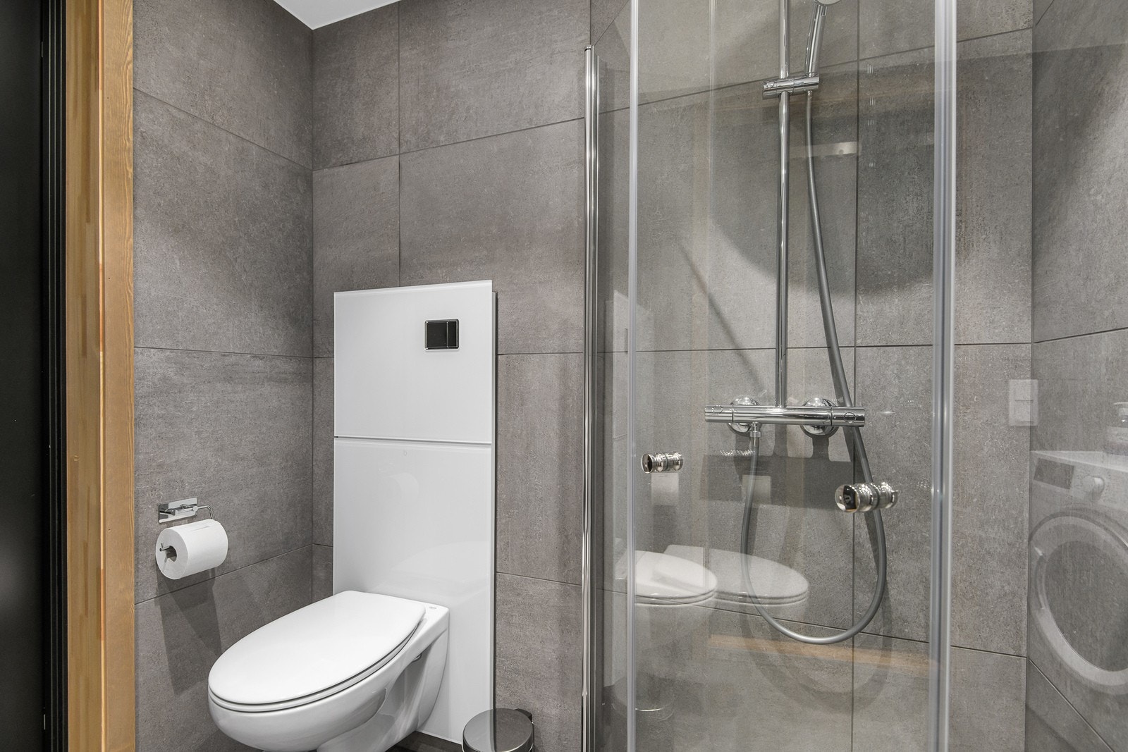 Badet er innredet med servant med underskap, speil med tilhørende belysning, dusjvegger, opplegg for vaskemaskin og vegghengt wc. Galleribilde