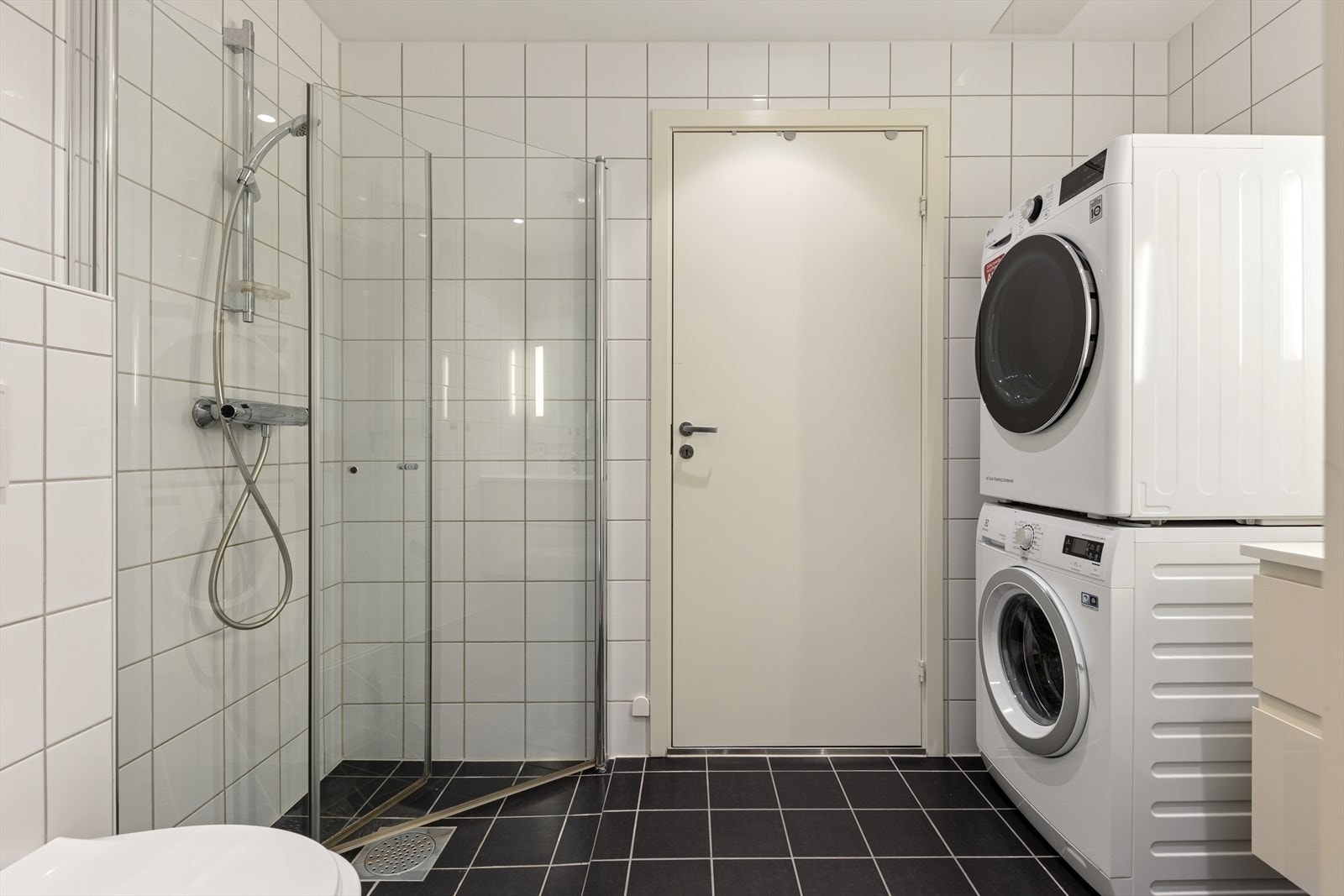 Badet har lys innredning, vegghengt toalett, dusjsone med dusjdører og opplegg for vaskemaskin og tørketrommel i høyden. Galleribilde