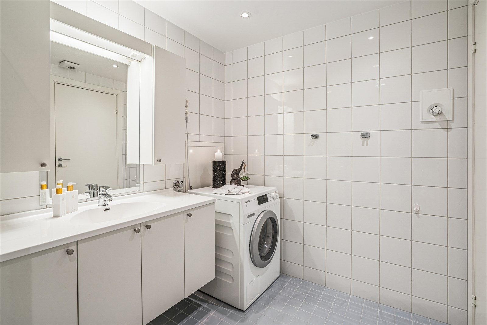 Badet er innredet med et vegghengt klosett, servant nedfelt i innredning og et dusjhjørne med svingbare glassdører. Det er også opplegg for vaskemaskin. Galleribilde