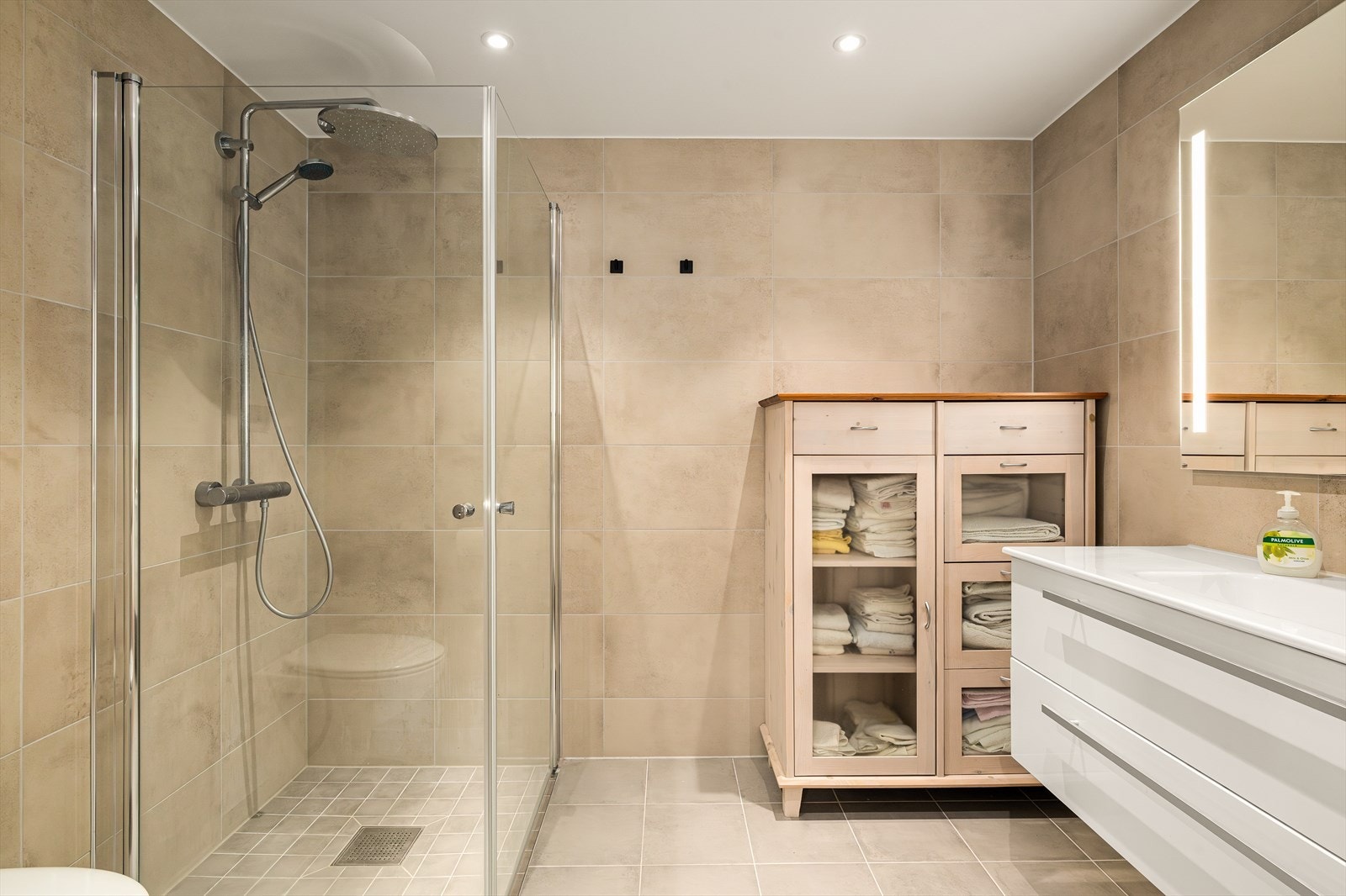 Rommet er innredet med servantskap og en elegant, nedfelt servant med praktisk enhåndsarmatur. Dusjen er plassert i en egen nisje med innfellbare glassdører som sparer plass og gir et rent uttrykk. Galleribilde