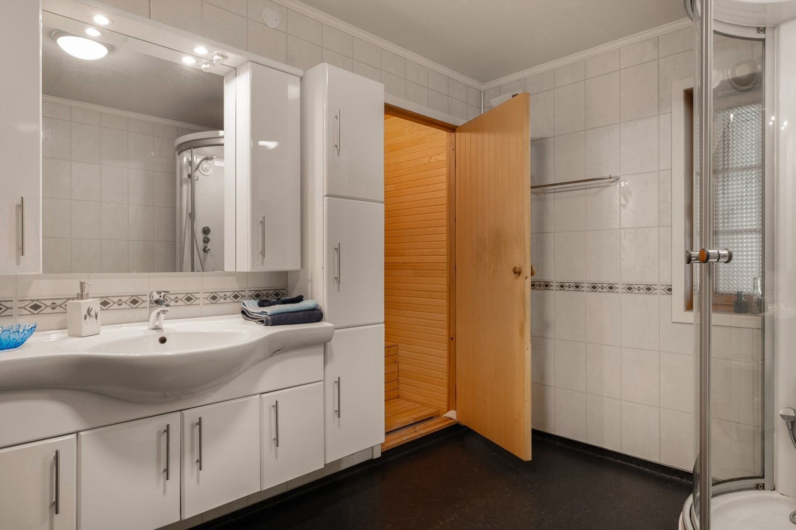 Badet er praktisk innredet med en romslig skapinnredning med heldekkende servant, dusjkabinett og gulvmontert wc. Galleribilde