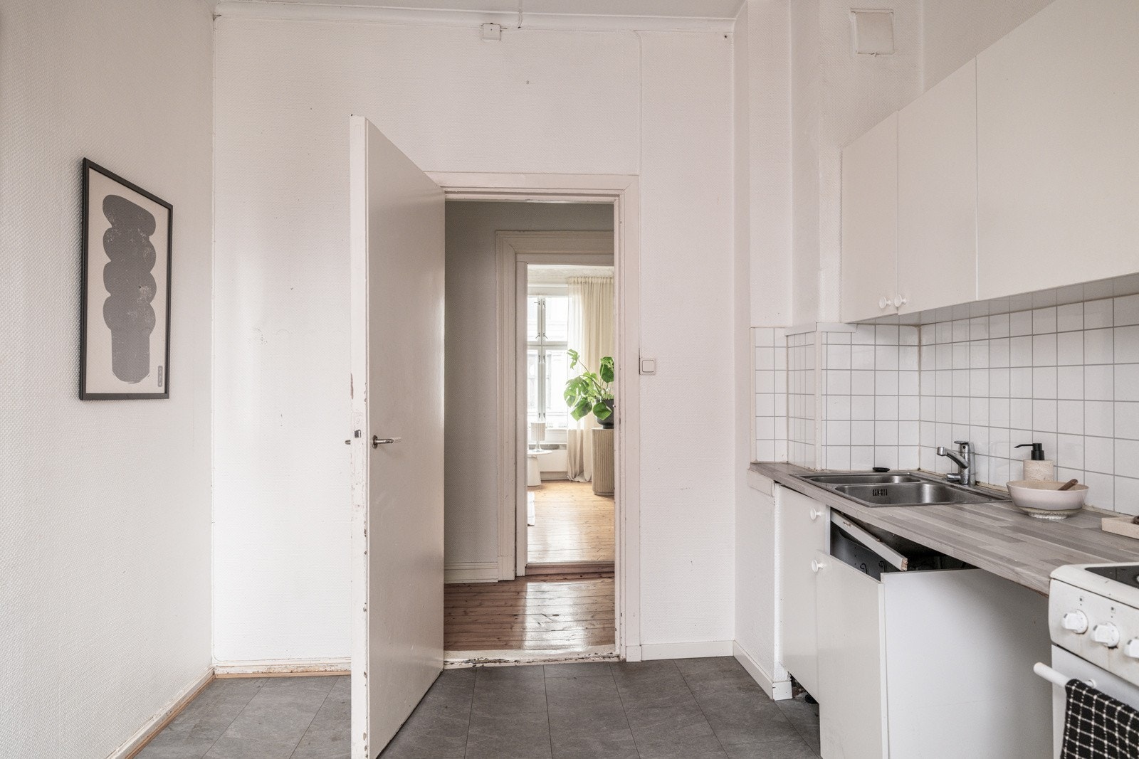 Eldre kjøkkeninnredning med behov for utskiftning. Innredningen har hvite glatte fronter, laminert benkeplate med nedfelt oppvaskkum og fliser i benkeryggen. Kjøkkenet er utstyrt med eldre komfyr og oppvaskmaskin Galleribilde