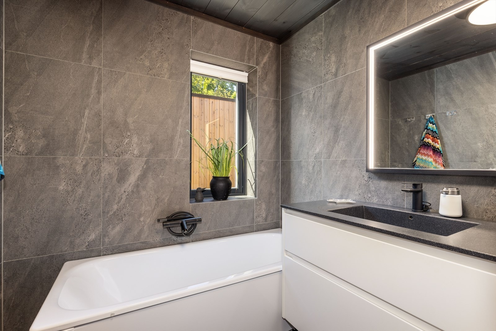 Badet er helfliset med store, moderne fliser og vannbåren varme i gulvet. Tidløs innredning med skuffer og heldekkende servant. Speilet på veggen gir et stilrent uttrykk kombinert med varm og behagelig belysning. Galleribilde