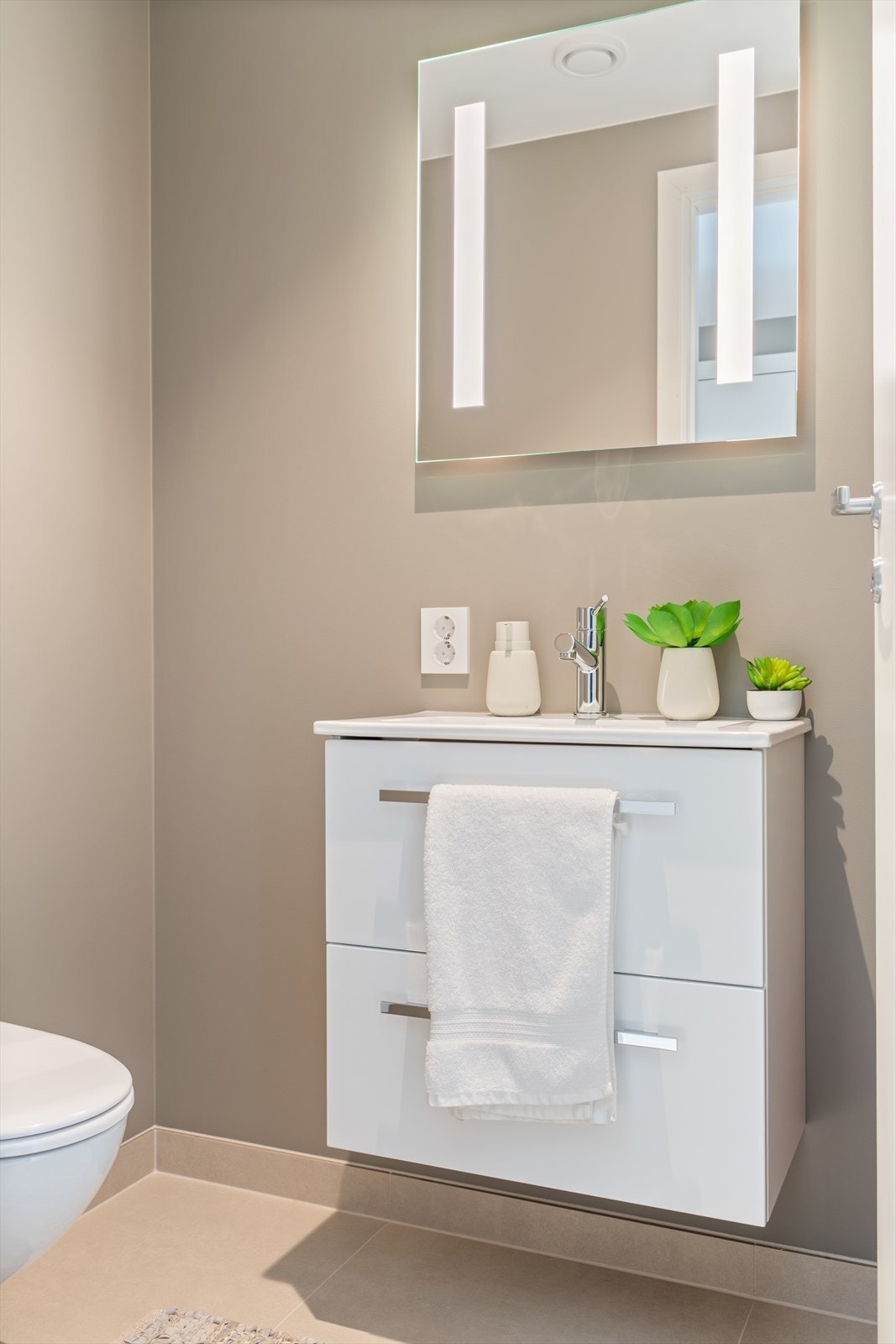 Lyst og pent toalettrom med fliser på gulv og malte vegger. Rommet er innredet med skuffeseksjon med servant og vegghengt toalett. Galleribilde