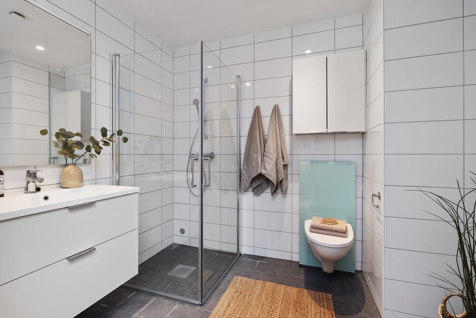 Pent flislagt bad med servantinnredning med heldekkende servant-topp, speil med belysning, dusjhjørne, vegghengt toalett og opplegg for vaskemaskin. Galleribilde