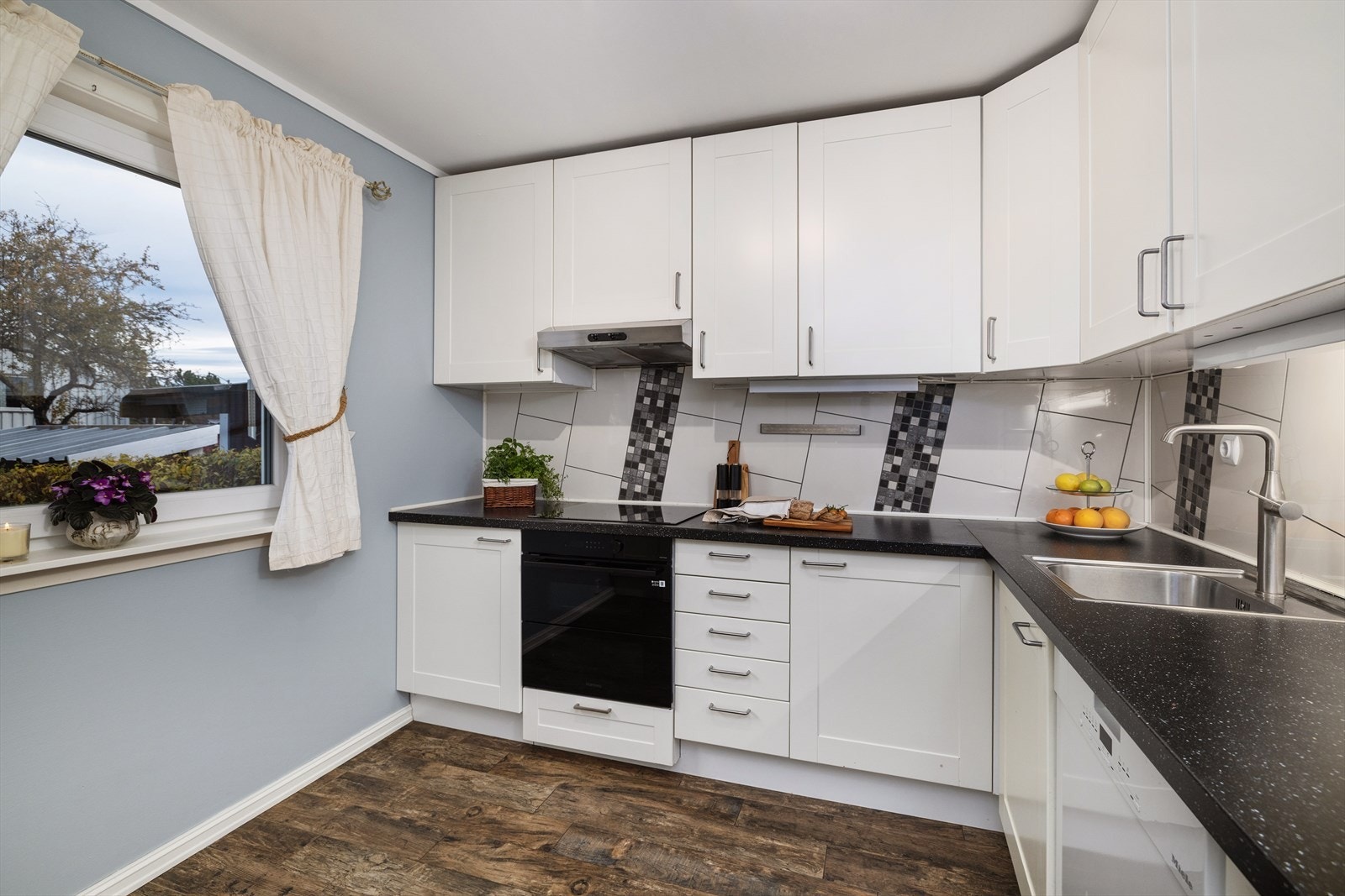 Laminat benkeplate. Flislagt bakvegg over benkeplate. Belysning under overskap. Integrerte hvitevarer som induksjon platetopp og stekeovn. Galleribilde