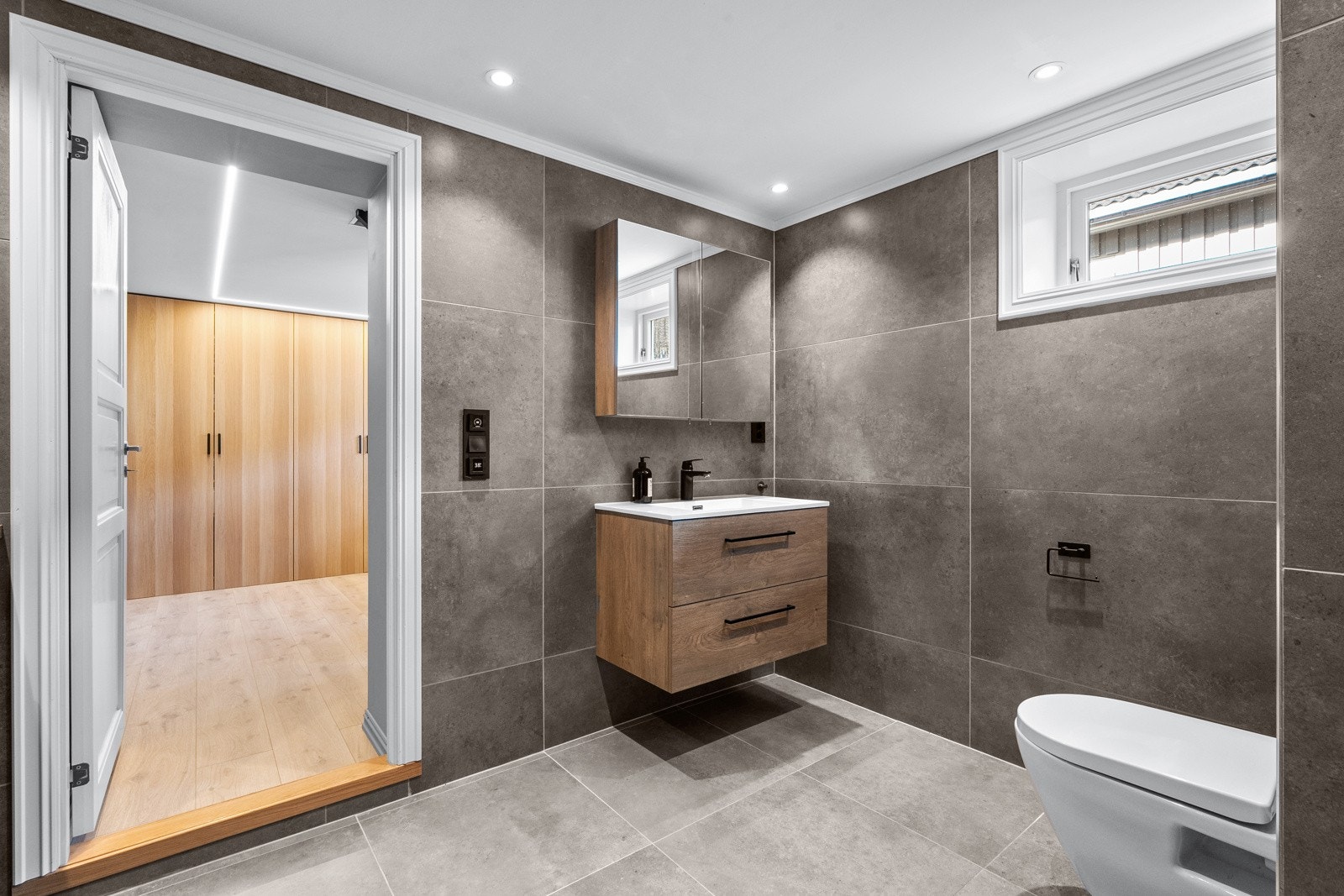 Rommet er innredet med vegghengt servantinnredning med skuffer, nedfelt servant, speilskap, vegghengt toalett og dusjhjørne. Galleribilde