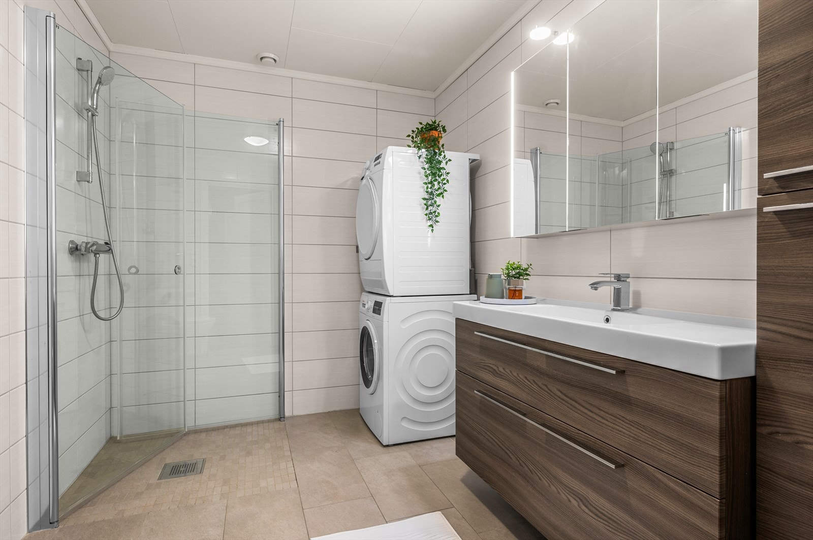 Leiligheten har et pent og moderne bad på 6 m², utstyrt med innfellbare dusjdører med dusjgarnityr på vegg, opplegg for vaskemaskin og moderne baderomsinnredning. Galleribilde