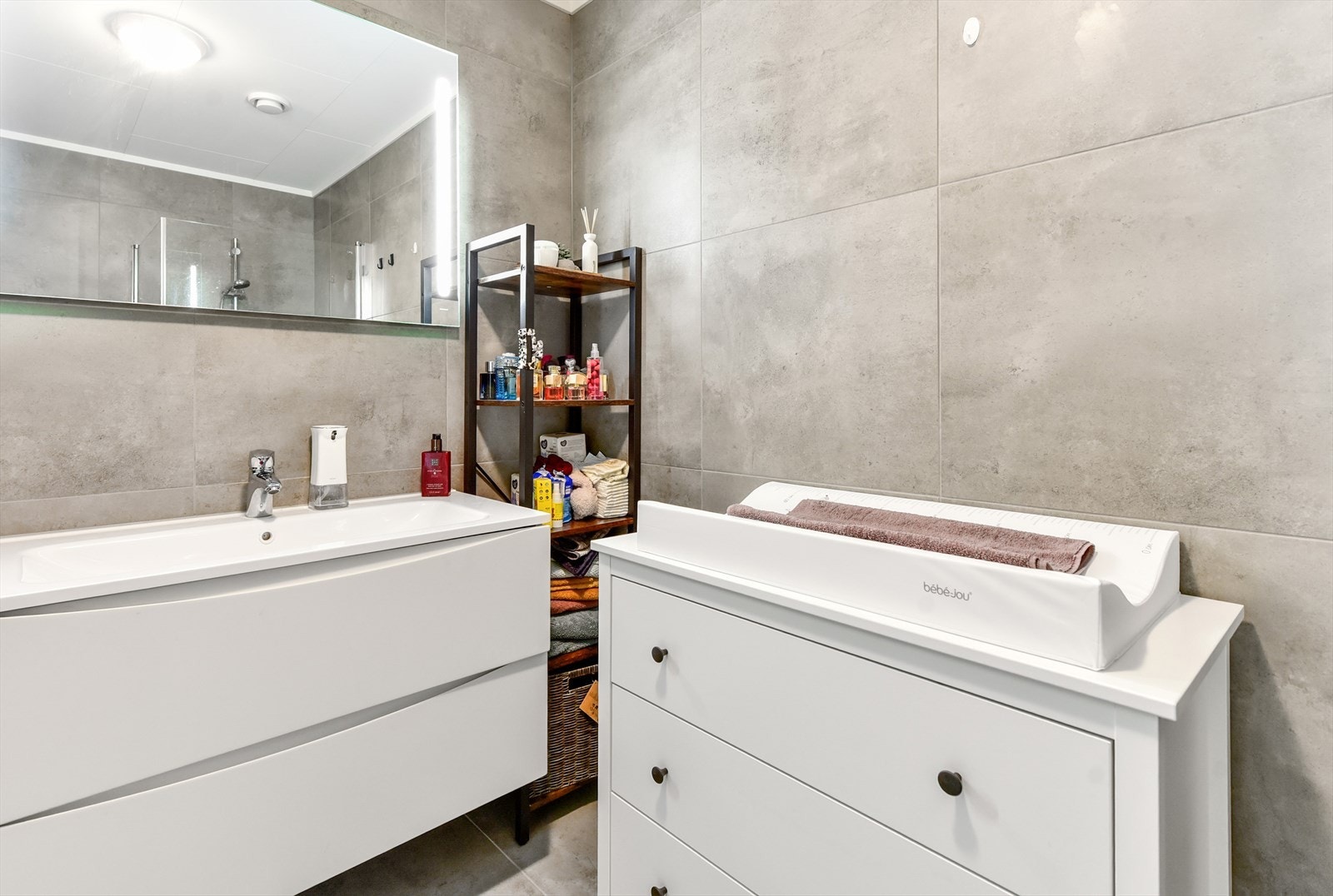 Lekkert, fliselagt bad med varmekabler i gulv, baderomsinnredning av type hvit glatt med heldekkende servant og to store skuffer, speil på vegg med lys, wc og dusjhjørne med dusjvegger i glass. Galleribilde