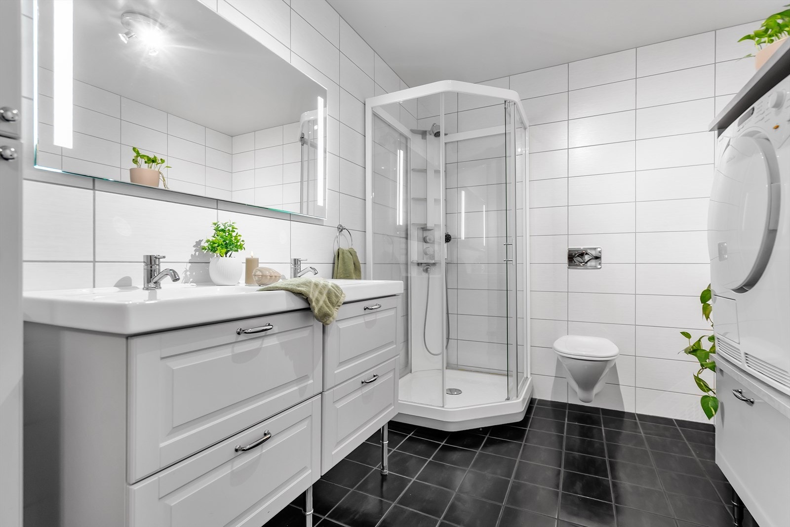 Delikat og moderne helfliset baderom som kombinerer funksjonalitet med stil. Rommet er innredet med dusjkabinett, vegghengt toalett og dobbel servant med underskap, samt et speil med integrert belysning som gir et lyst og elegant uttrykk. Galleribilde