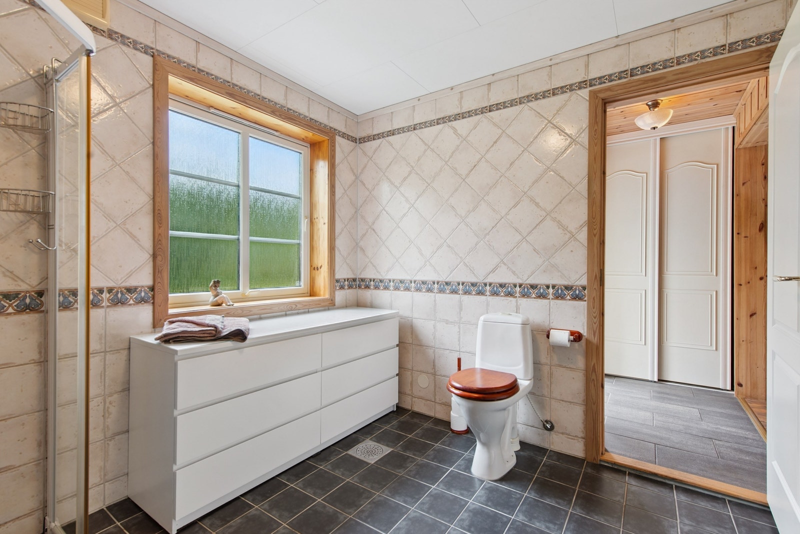 Badet er innredet med servantskap, overskap med speil, en dusjnisje i glass og metall, og et gulvmontert toalett. Oppvarming skjer via vannbåren gulvvarme. Galleribilde