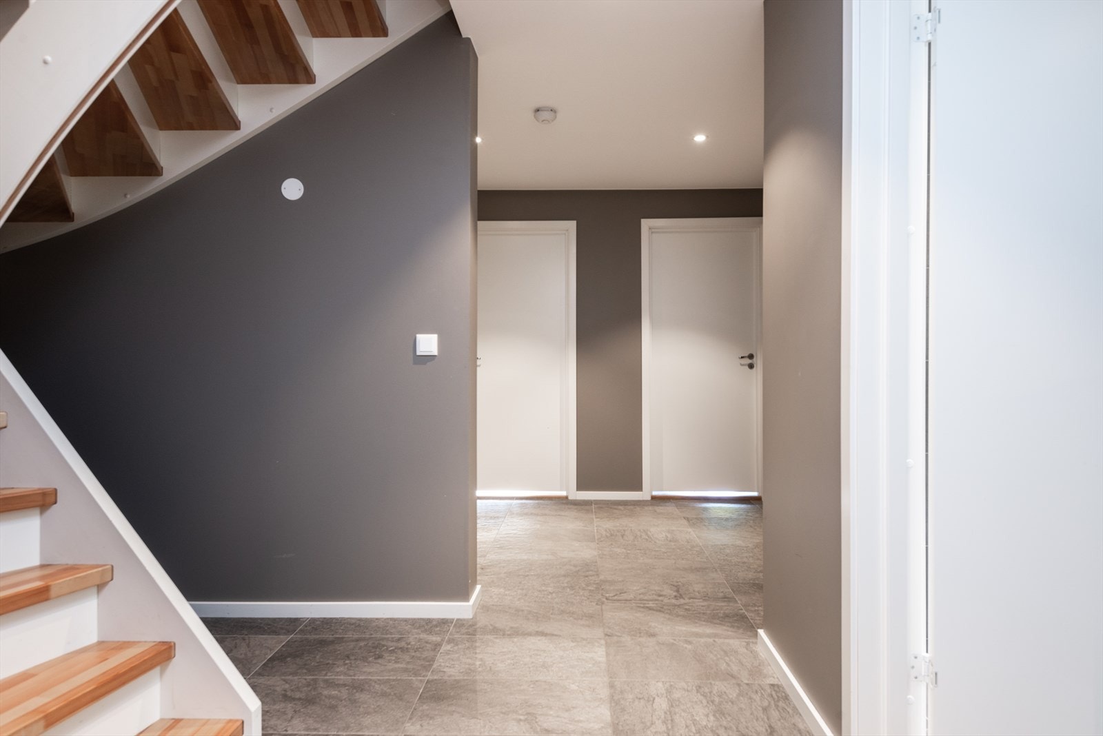 Flislagt gulv i entréen gir et solid og enkelt vedlikeholdt underlag. Trappen har en moderne design med tretrinn som gir en varm kontrast til de grå veggene. Galleribilde