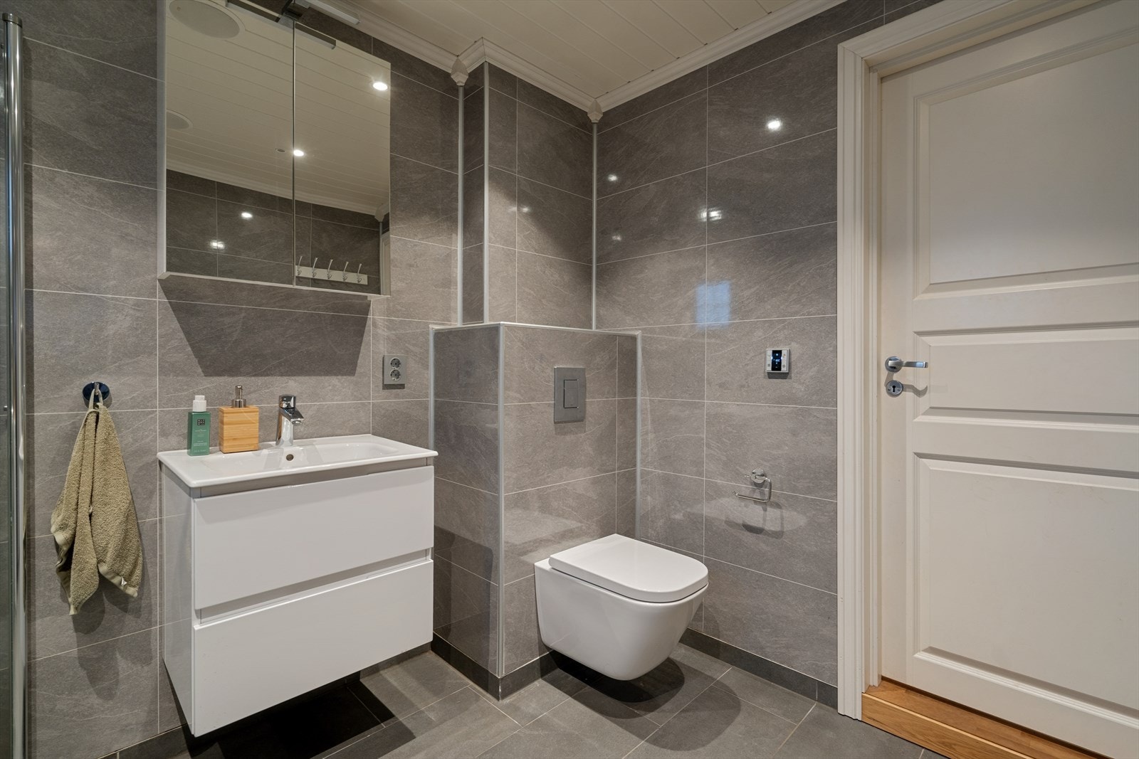 Baderommet fra 2018, fremstår moderne med flislagt gulv og elektriske varmekabler, baderomsplater på vegger og paneltak med downlights. Galleribilde