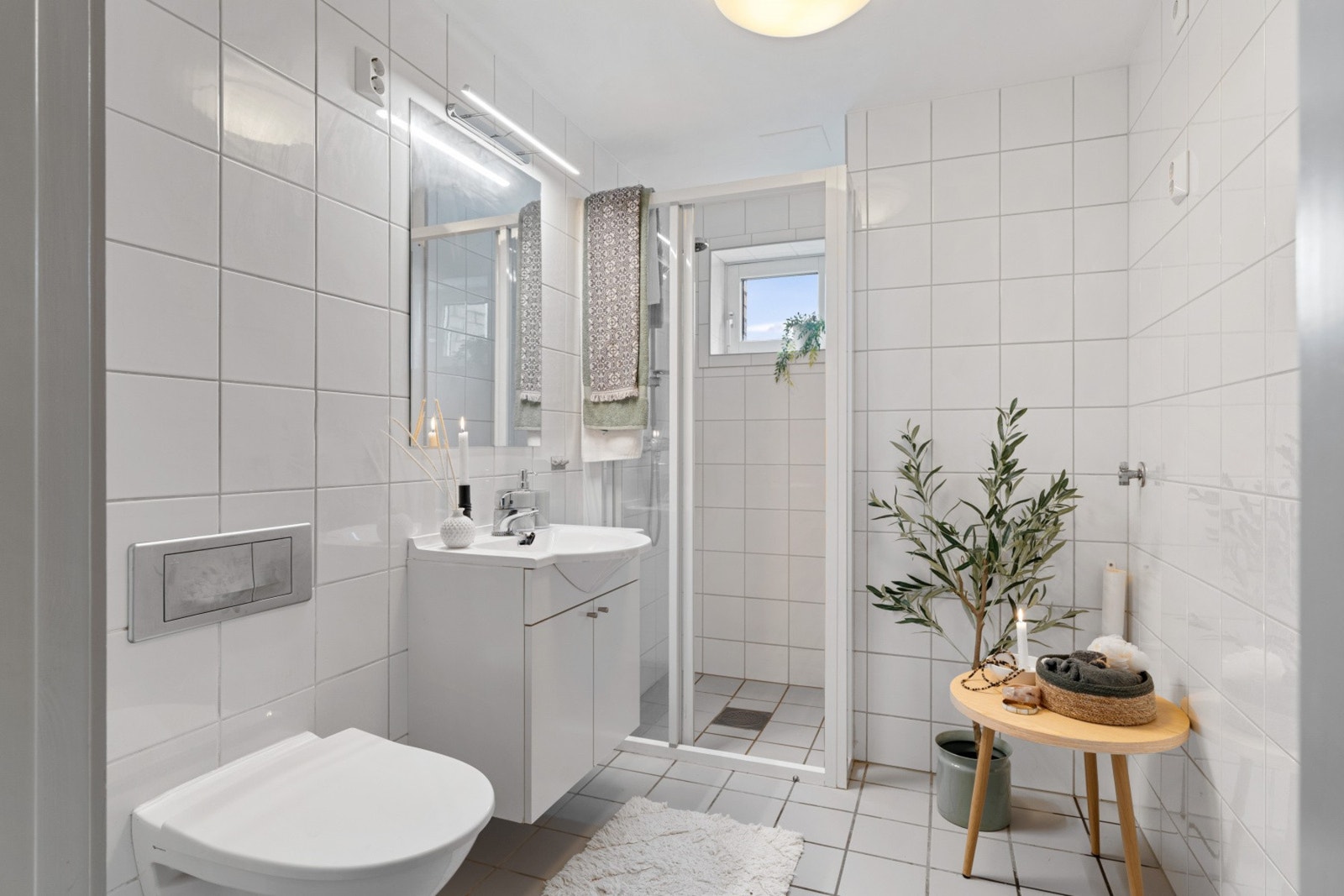 Lyst baderom med vegghengt toalett, dusjsone, servant med underskap og speil. Galleribilde