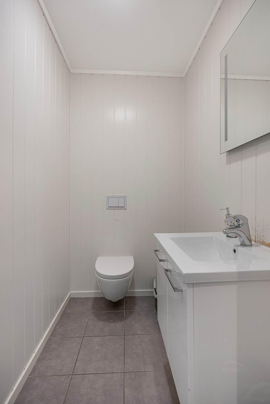 WC-rommet har flislagt gulv med varmekabler, vegghengt toalett, et hvitt servantskap og et speil med lys integrert. Galleribilde