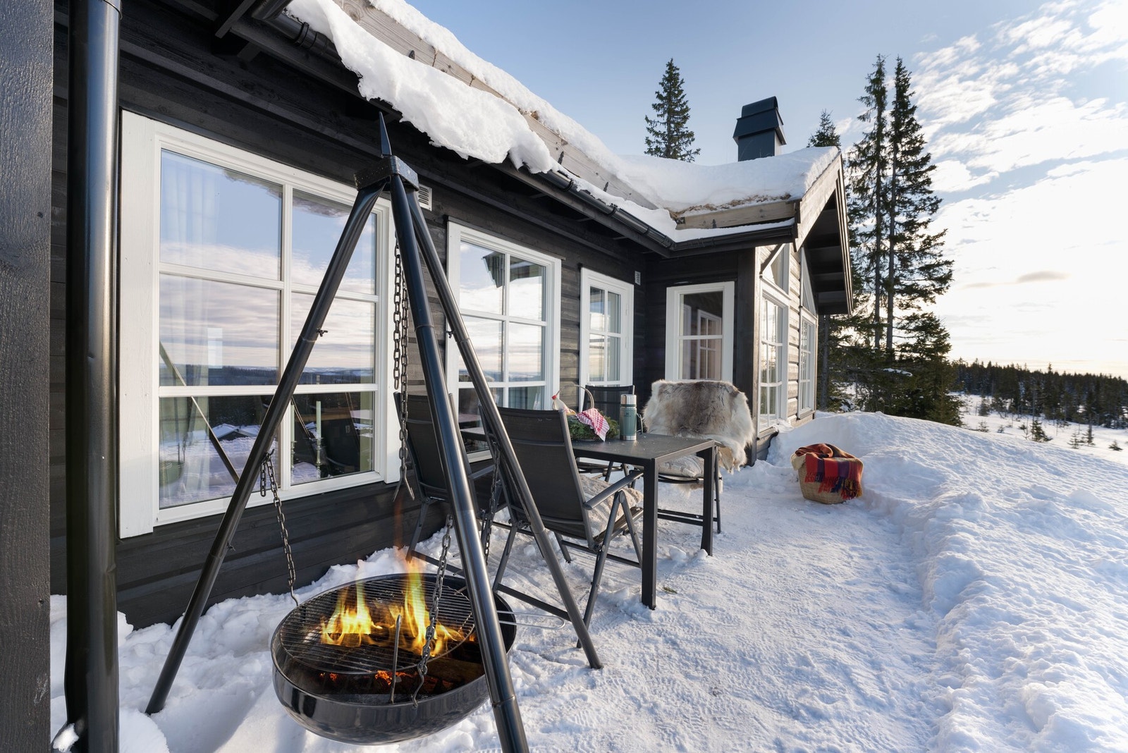 Solrik terrasse med sitteplasser og bålpanne. Hytta har store vinduer som slipper inn mye lys. Flott utsikt over snødekt landskap og skog. Galleribilde