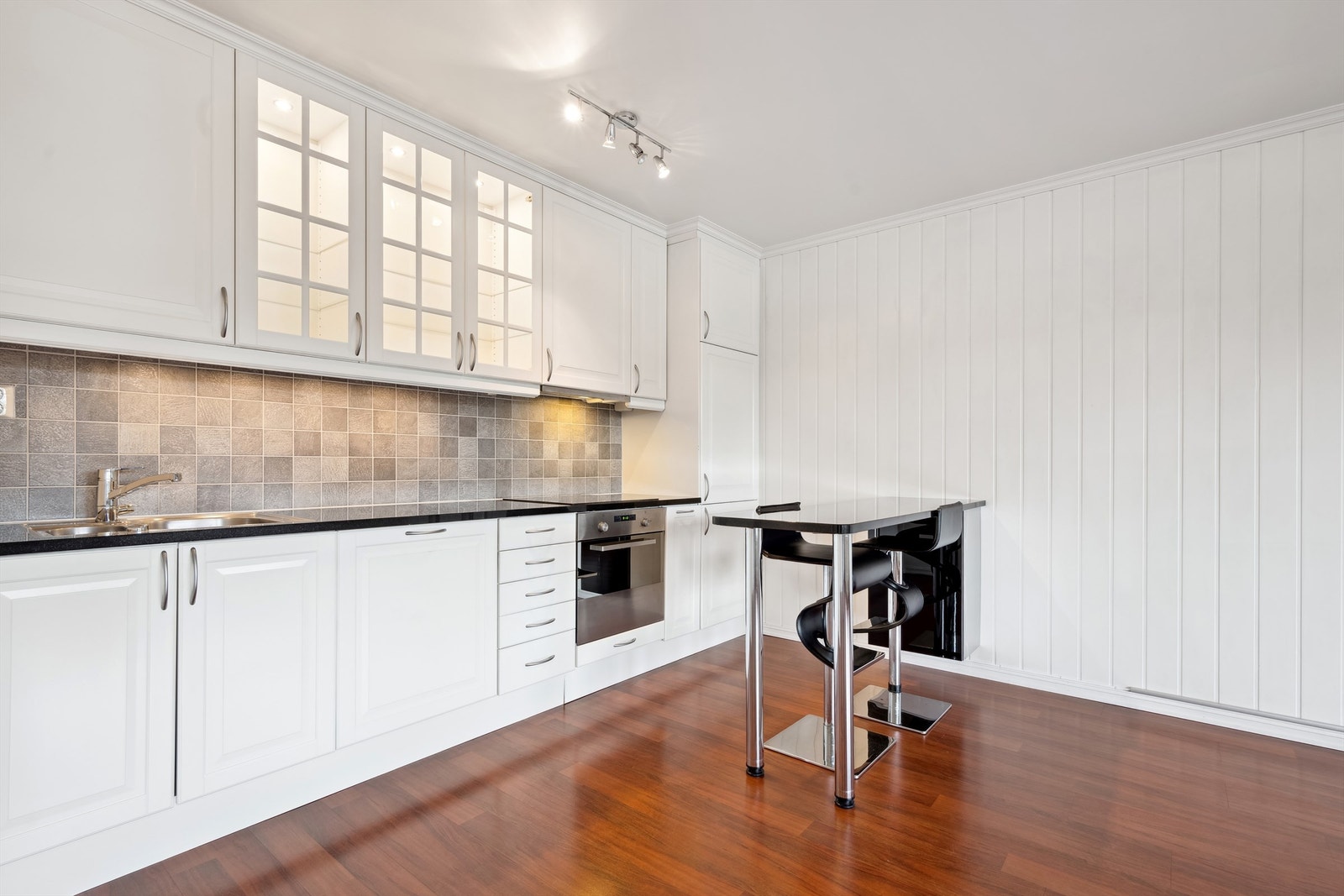 Pent kjøkken med innredning i hvite profilerte fronter, benkeplate av granitt i sort og integrerte hvitevarer Galleribilde