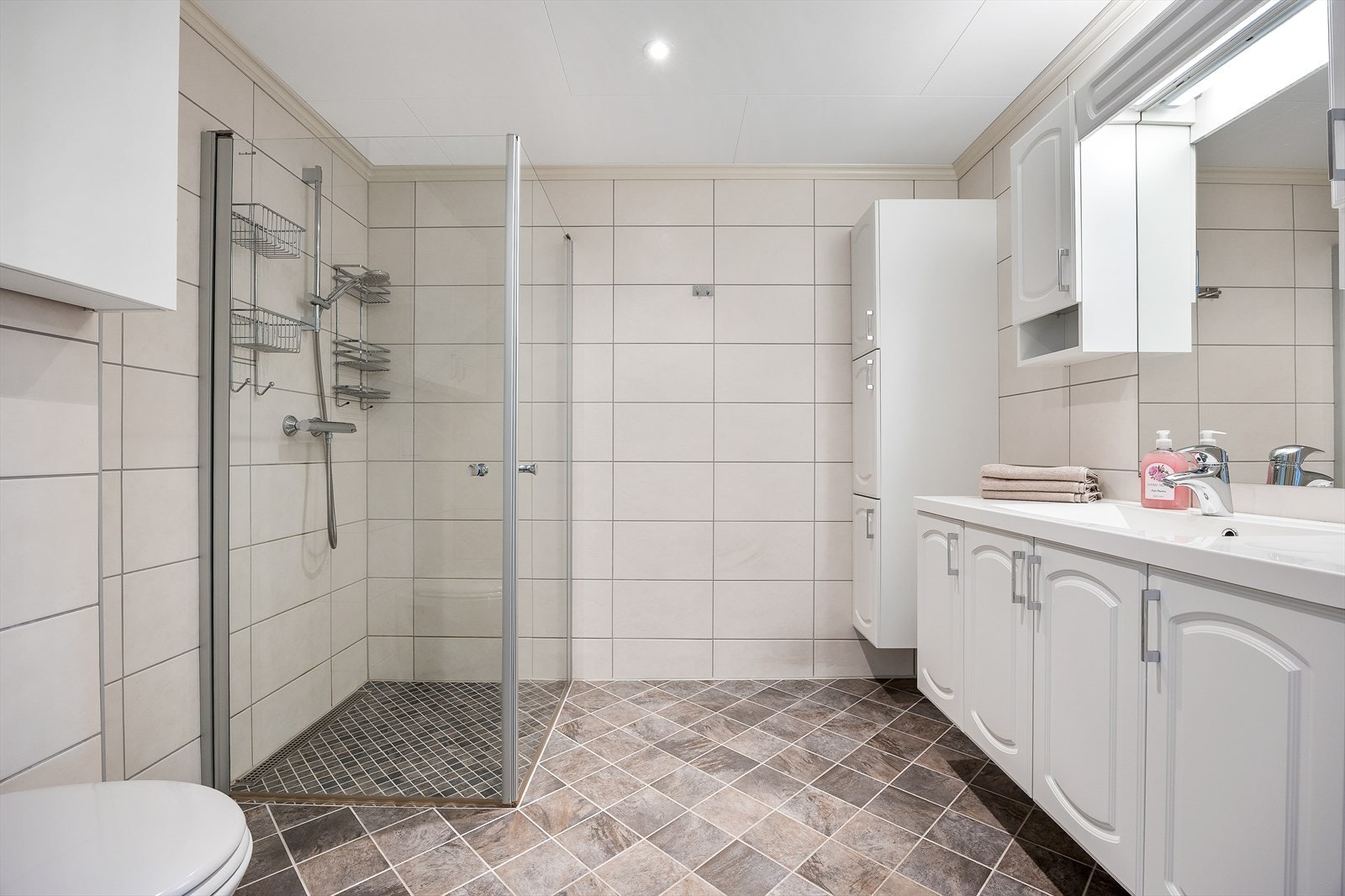 Baderom med flislagte gulv og vegger. Badet er utstyrt med vegghengt toalett, servant med skap og dusjnisje med innfellbare glassdører. Galleribilde