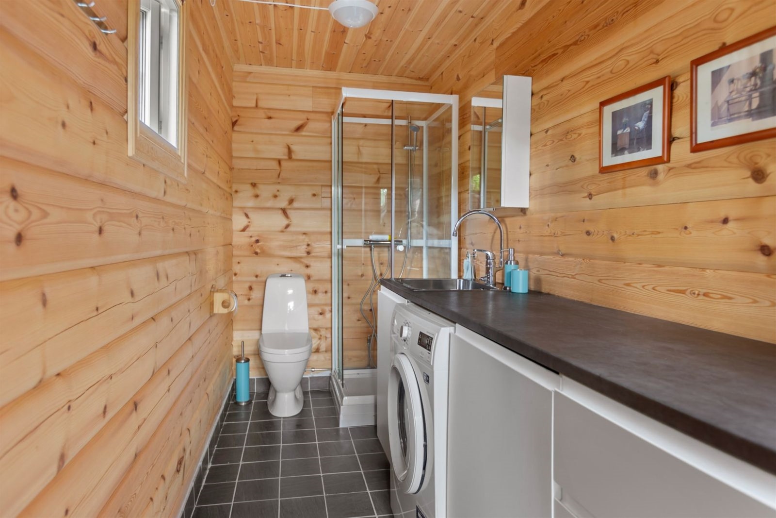 Bad i garasje som er innredet med gulvstående toalett, dusjkabinett, vaskekum og benkeplate med plass til vaskemaskin. Galleribilde