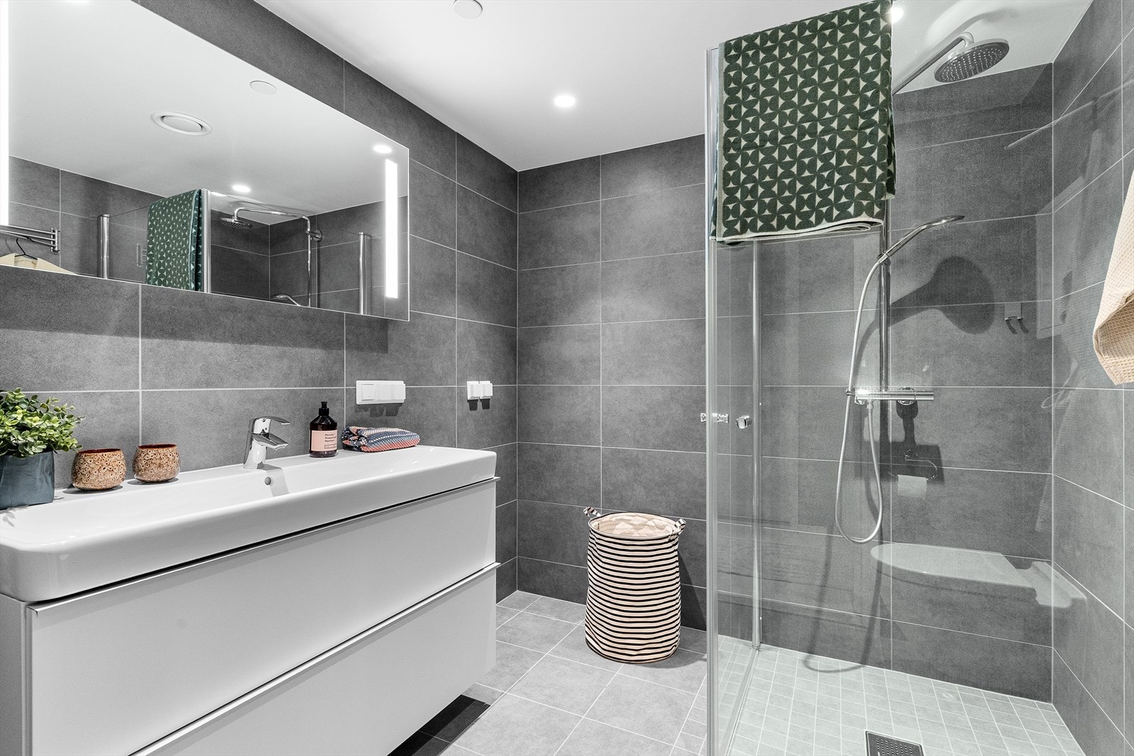 Baderomsinnredningen med heldekkende nedfelt servant og rom for oppbevaring i skuffeseksjon og speil med belysning. Videre har badet dusjhjørne med innfellbare dusjvegger og vegghengt toalett. Galleribilde