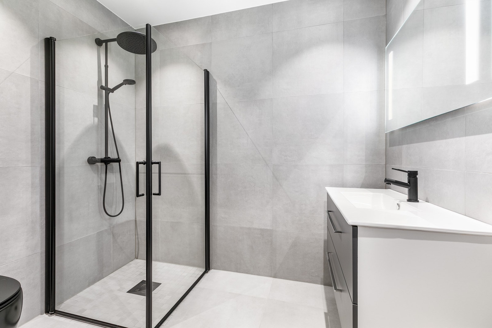 Badenes gulv og vegger er helhetlig utført med like fliser av storformat, dusjsonens gulv med mosaikk. Galleribilde