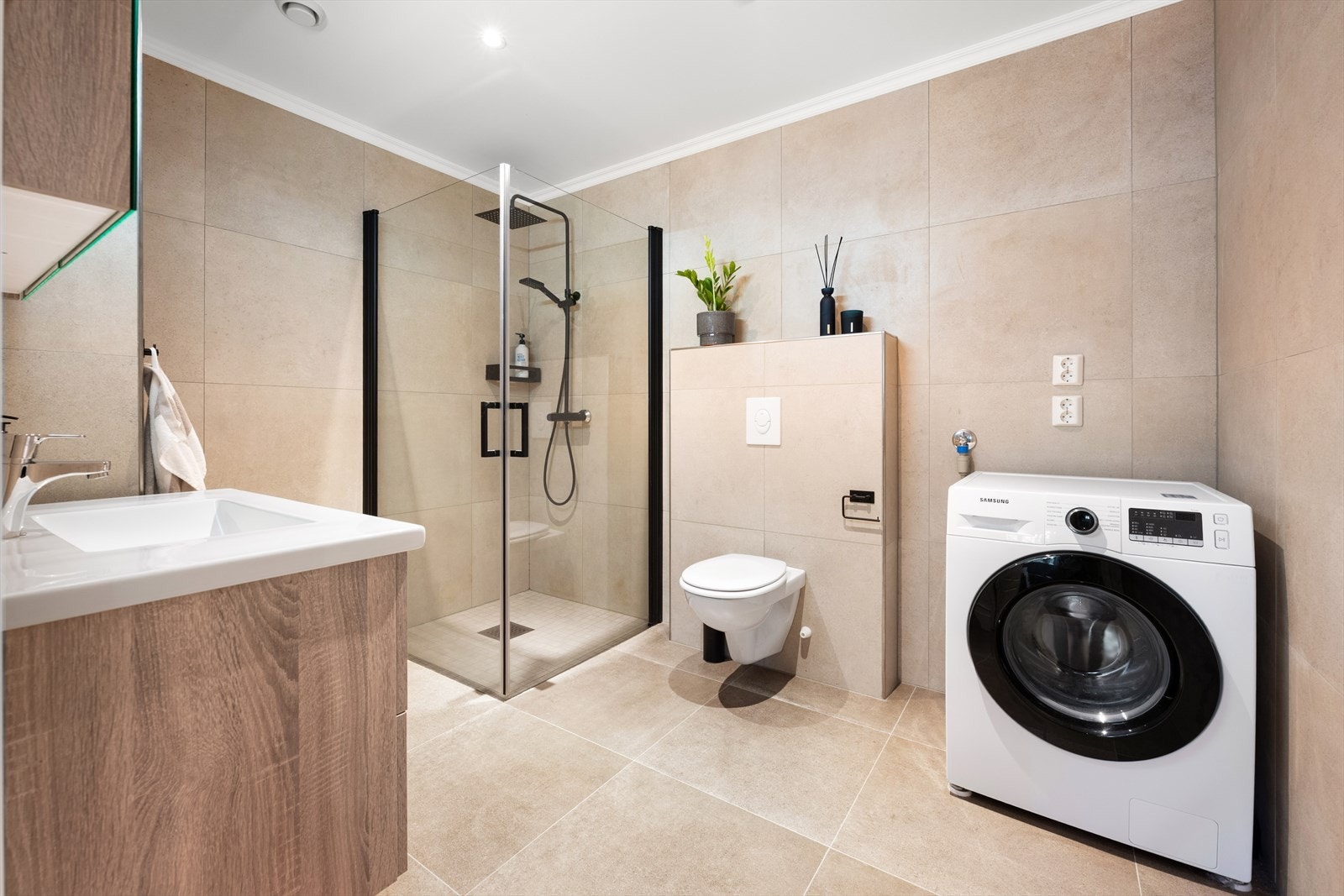 Baderomsinnredningen har heldekkende nedfelt servant og rom for oppbevaring i skuffeseksjon og speil med belysning. Videre har badet dusjhjørne med innfellbare dusjvegger og vegghengt toalett. Rommet har opplegg for vaskemaskin. Galleribilde