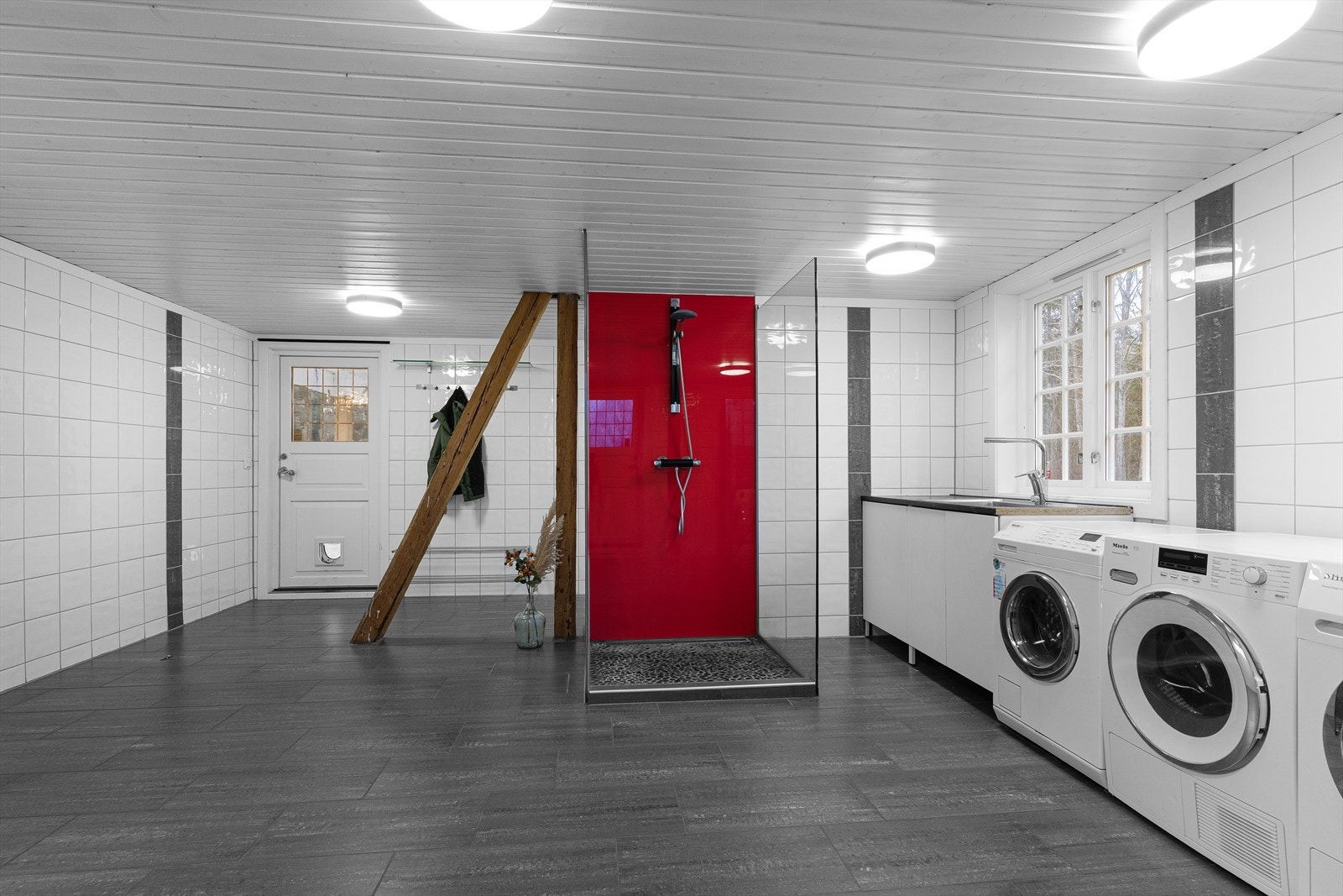 Det er flislagt gulv og vegger samt elektrisk gulvvarme. Dette er et meget stort rom inndelt i to soner. Den ene sonen har opplegg for vaskemaskiner og tørketrommel samt benk og vaskekum. Midt i rommet er det dusj med glassvegger. Galleribilde