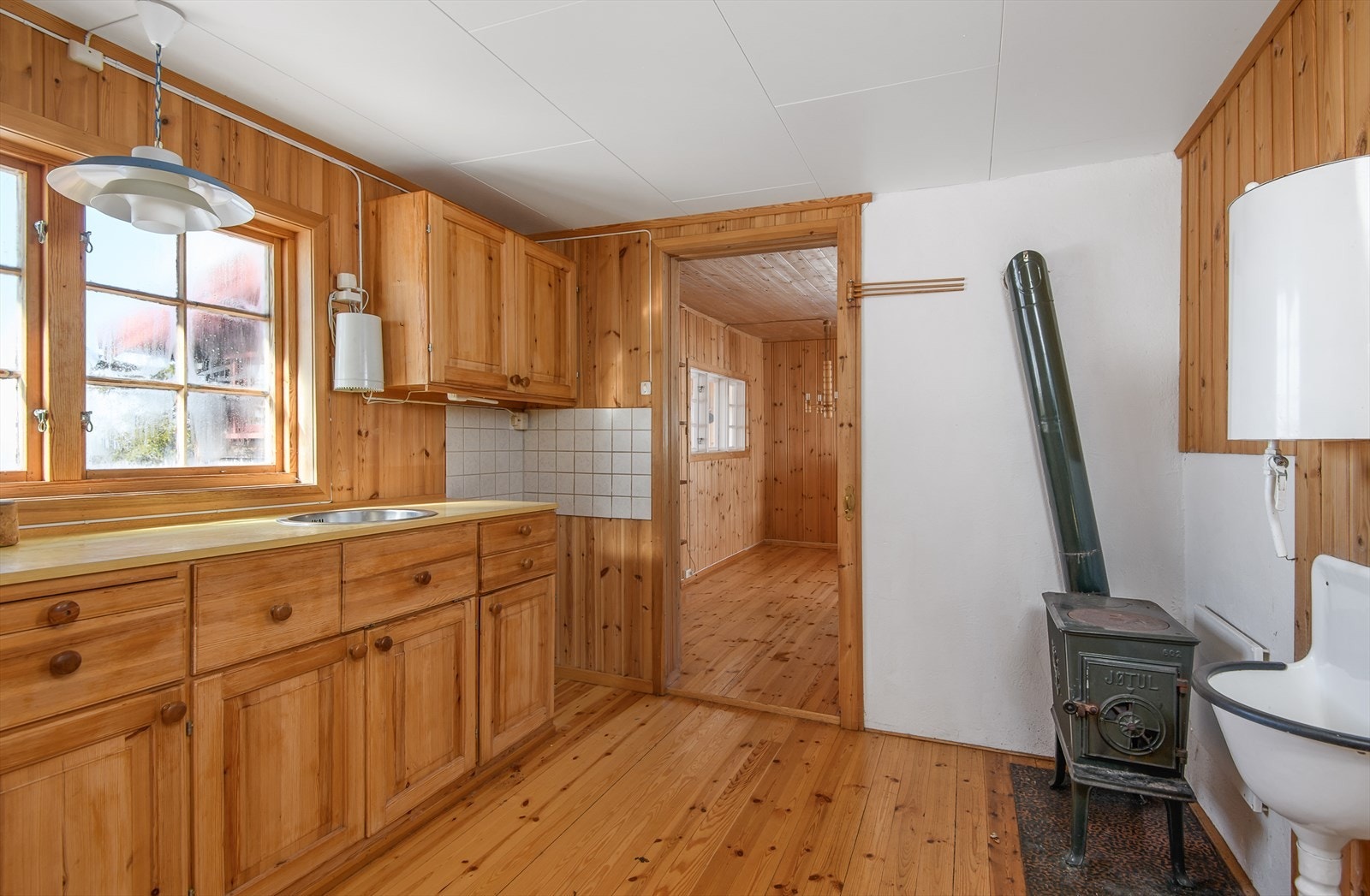 Kjøkkenet har enkel innredning langs den ene veggen. Trehvite fronter. Laminat benkeplate. Fint lys inn gjennom vindu over kjøkkenbenken. Galleribilde