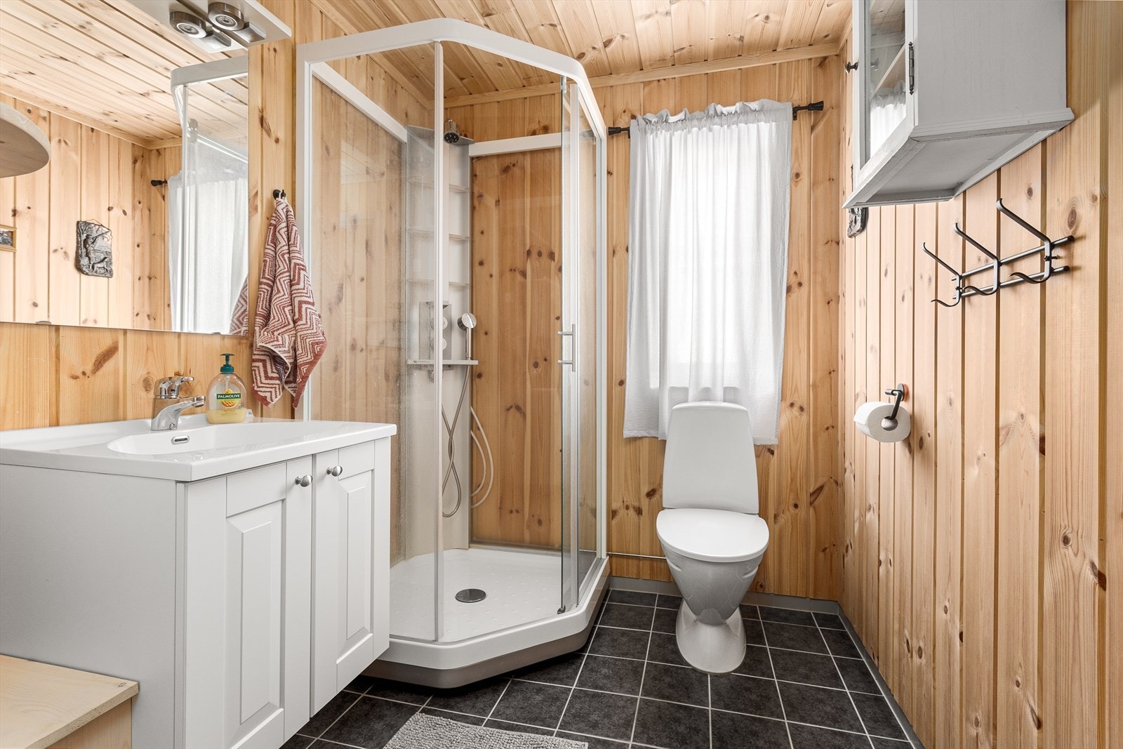 Badet har panel på veggene og flislagt gulv med elektriske varmekabler. Innredningen består av nedfelt servant, toalett og dusjkabinett. Galleribilde