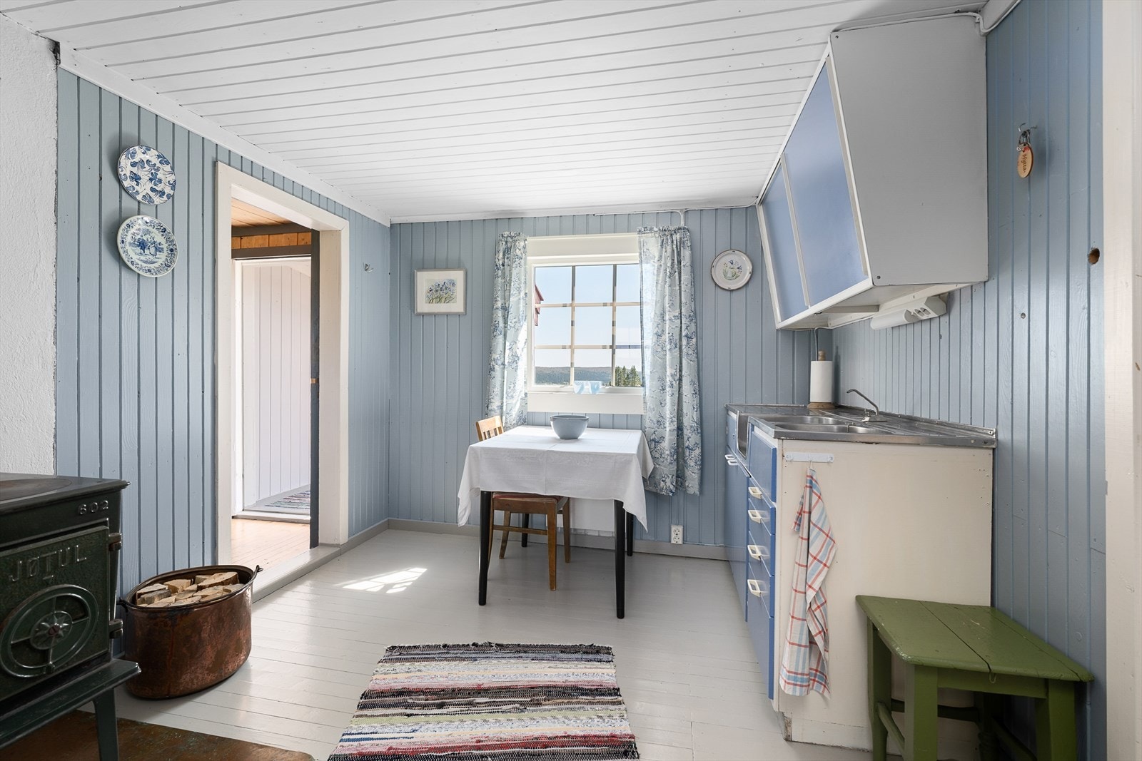 Den gamle panelen på veggene på kjøkkenet er malt i en dus og delikat blåtone. Vedovn for god oppvarming i rommet. Galleribilde