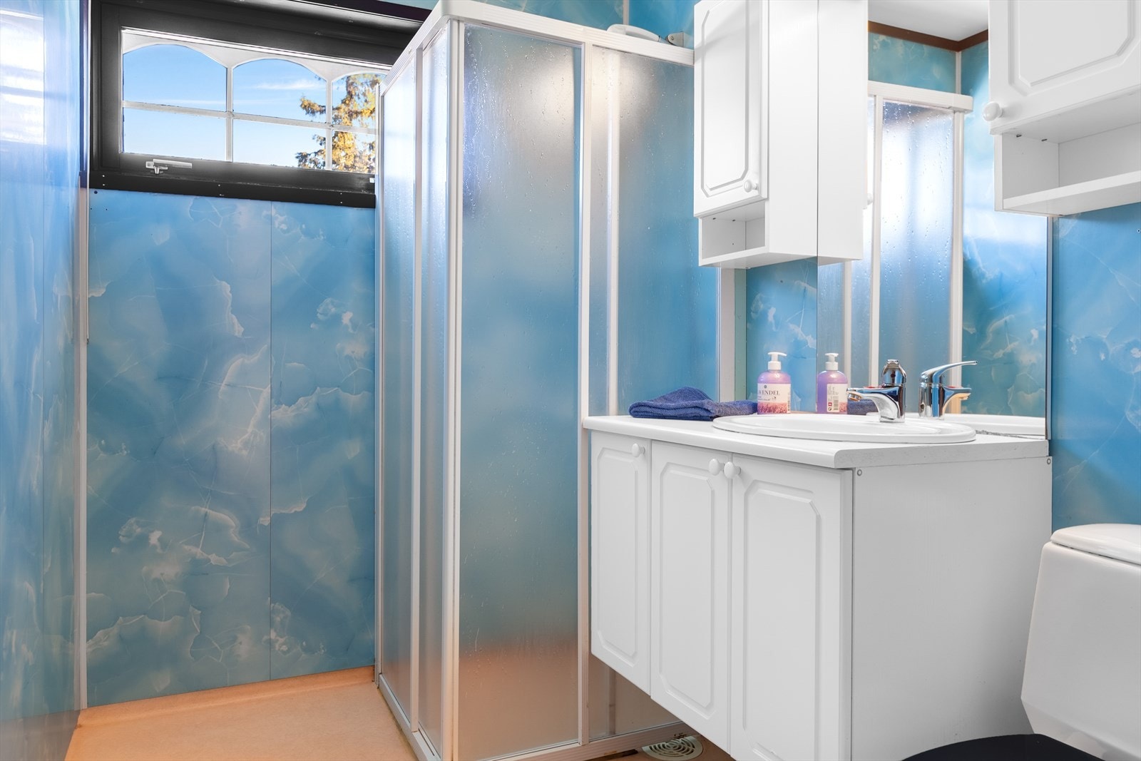 Badet er fra byggeår med belegg på gulv og baderomsplater på vegg. Her er det lys baderomsinnredning, vegghengt dusj med glassvegger og frittstående toalett. Badet opplyses å ha behov for totalrenovering. Galleribilde