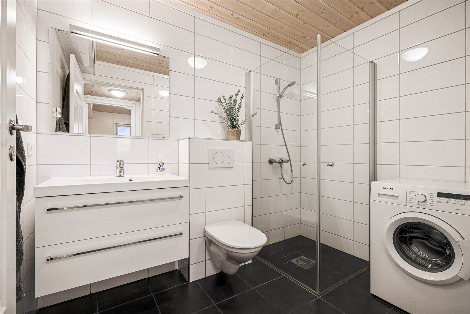 Badet inneholder dusjhjørne, vegghengt toalett, servant med skuffer, og opplegg for vaskemaskin. Galleribilde