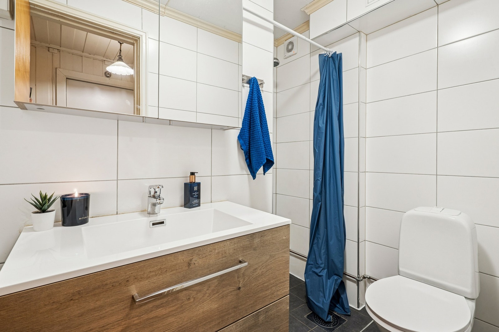 Dusjnisje med dusjforheng og dusjarmatur. Gulvmontert toalett. Vannrør av kobber. Synlige avløpsrør av plast. Plastsluk i gulv. Mekanisk avtrekksvifte. Galleribilde