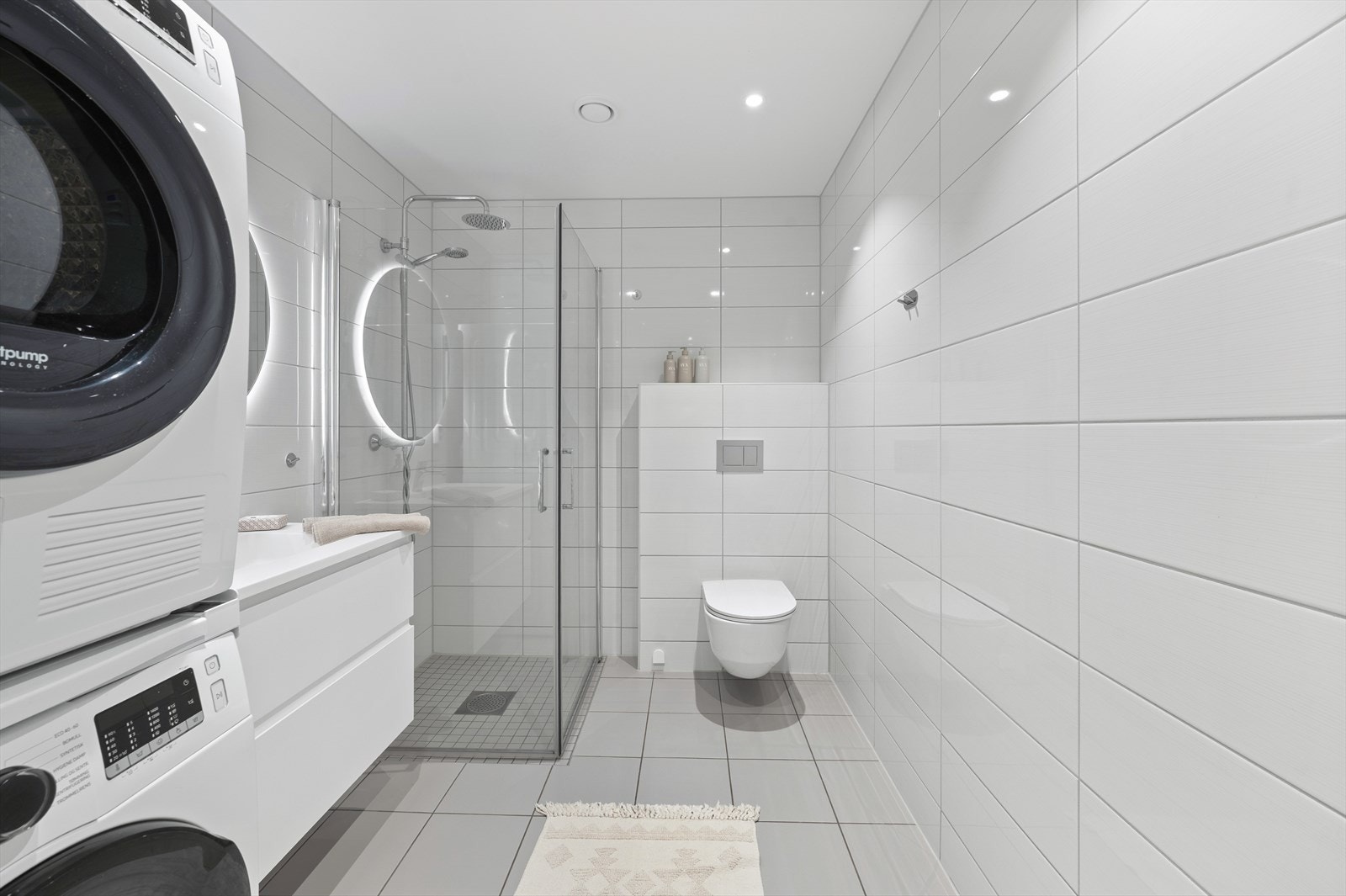 Badet er innredet med heldekkende nedfelt servant med rom for oppbevaring i skuffer, rundt speil med led lys, vegghengt toalett og dusjhjørne med innfellbare dusjvegger. Opplegg for vaskemaskin og tørketrommel. Galleribilde