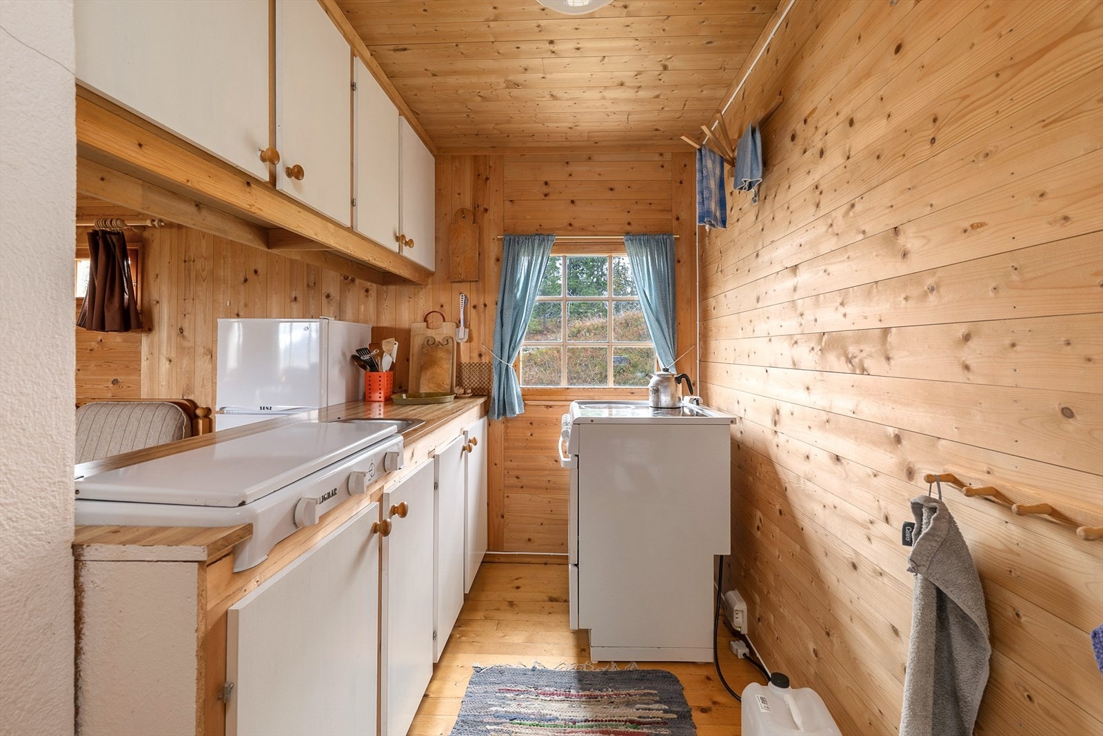 Kjøkkeninnredningen har hvite, glatte fronter. Over-og underskap for god oppbevaring. Propan kokeapparat plassert på benk. Komfyr plassert ved vinduet. Galleribilde