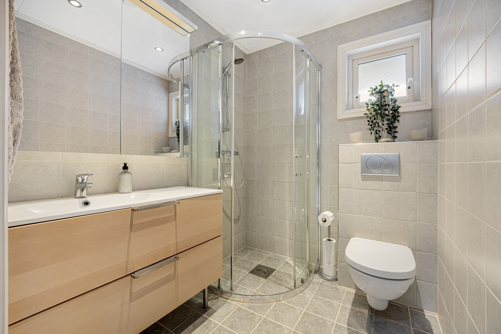 Det helflisede badet i 1. etasje har vegghengt toalett, dusjhjørne med skyvedører av herdet glass og varme lagt i gulvet. Innredningen har heldekkende servant over skuffeseksjon og speilskap med belysning i overkant. Galleribilde