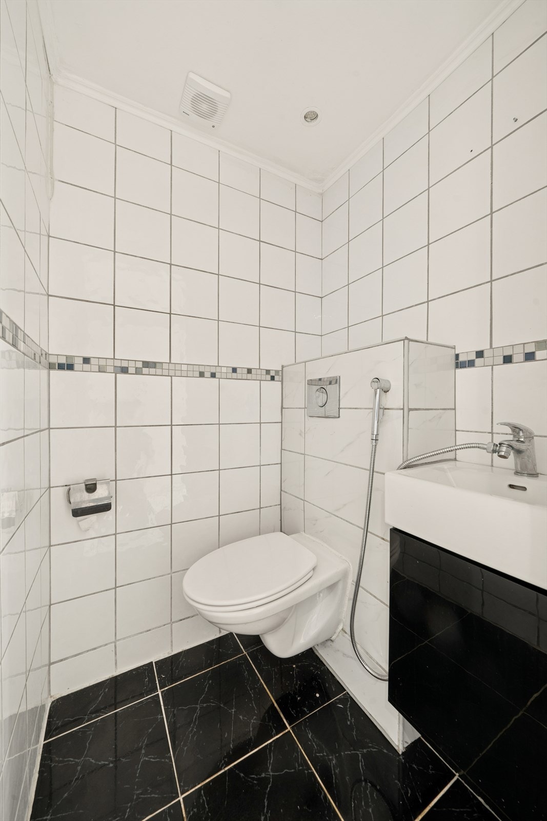 Et separat og praktisk toalettrom med fliser på gulv og vegger. Rommet er utstyrt med et moderne, vegghengt toalett og en servant. Galleribilde