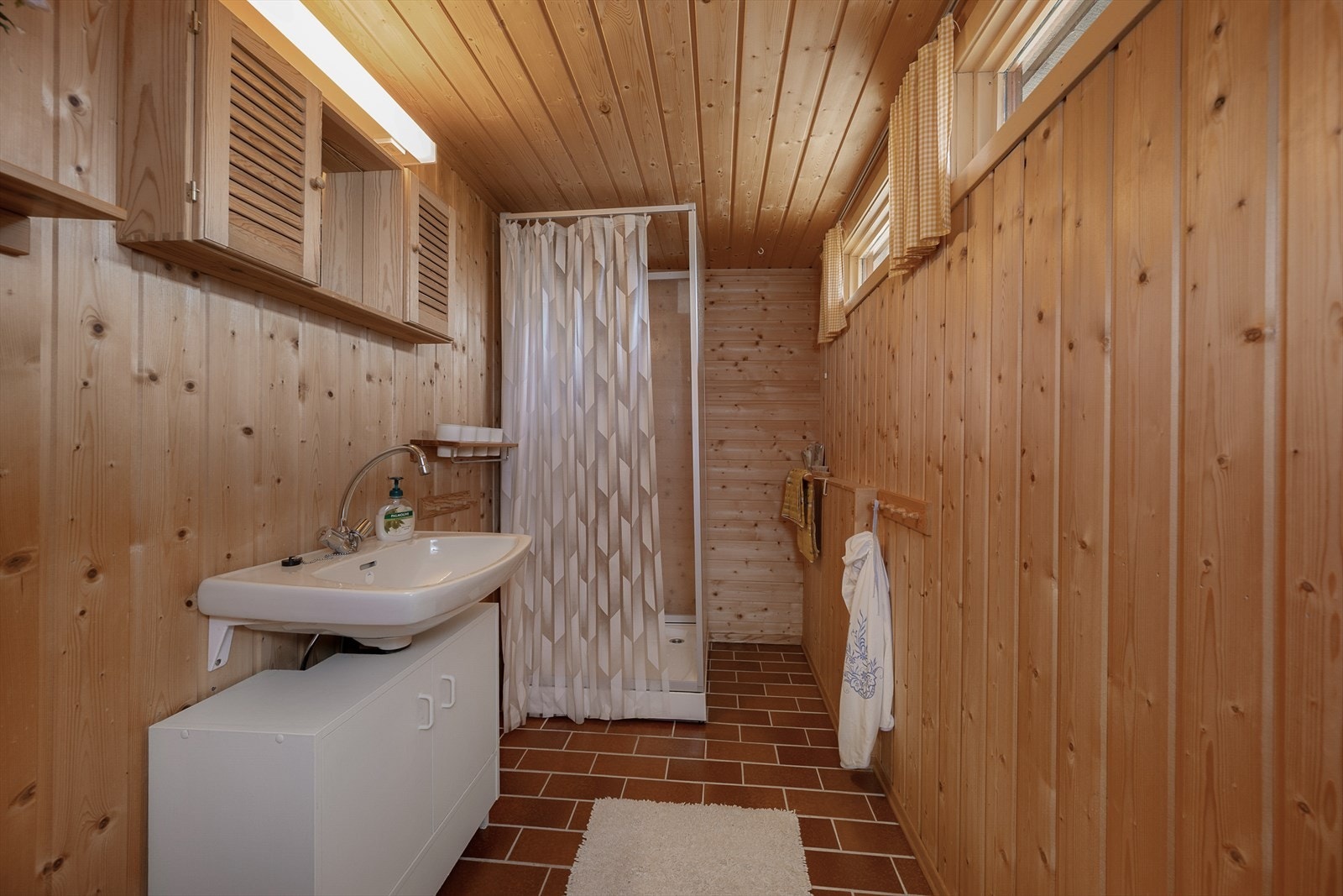 Hyttas baderom har enkel servant med skap under og dusjkabinett. Panelte vegger. Fliser på gulv. Galleribilde