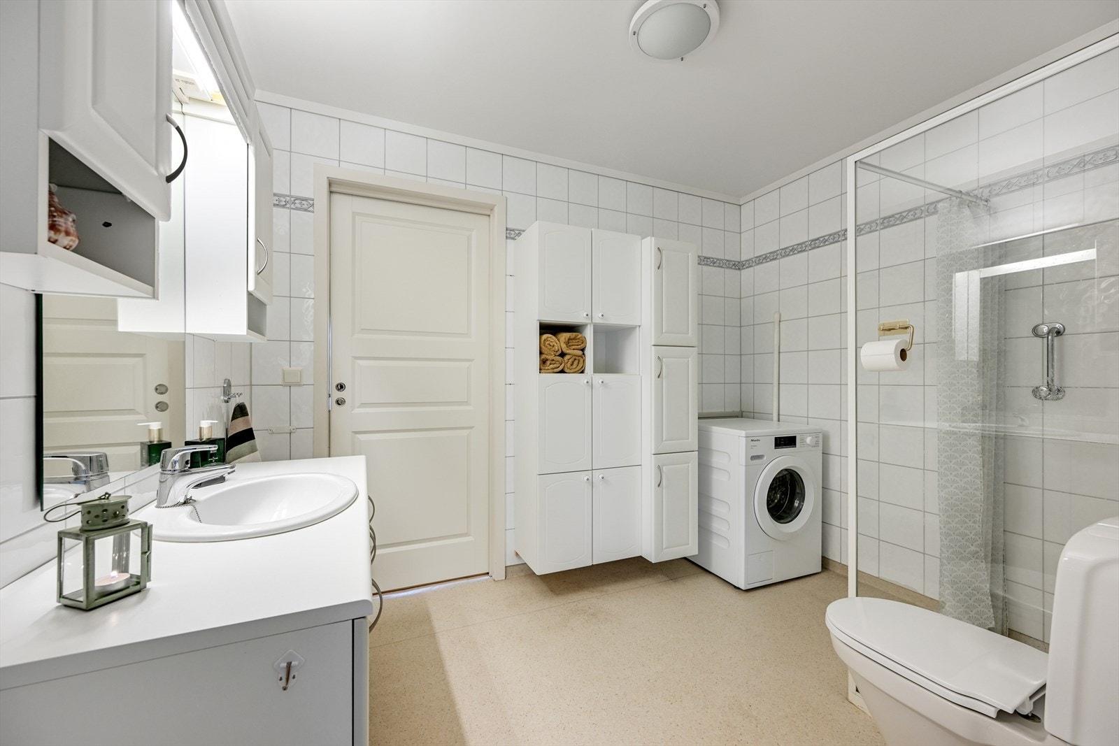 Romslig bad med belegg på gulv, og flis på vegg. Servant i skapinnredning, høyskap på vegg, dusj på vegg med glassvegg, toalett og opplegg for vaskemaskin. Galleribilde