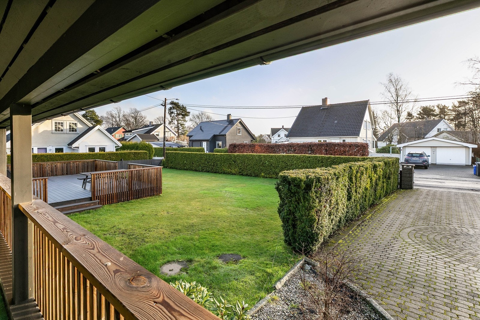Utsyn fra kjøkkenets terrasse til hagen og innkjørselen. Innsyn fra gaten begrenses av den tettvoksende hekken. Galleribilde