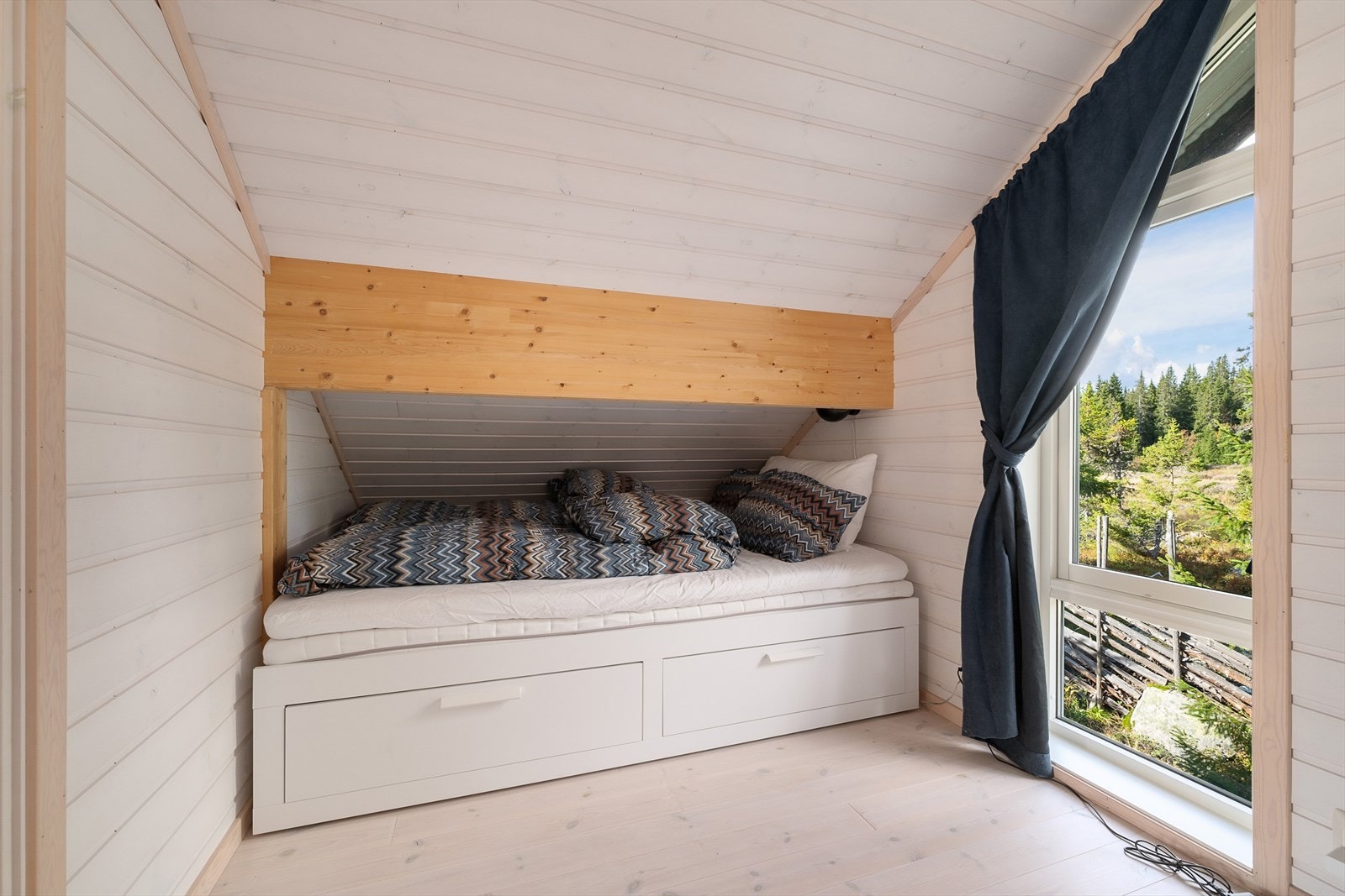 Soverom med seng under skråtaket med praktisk oppbevaring. Fin utsikt gjennom soveromsvinduet. Galleribilde