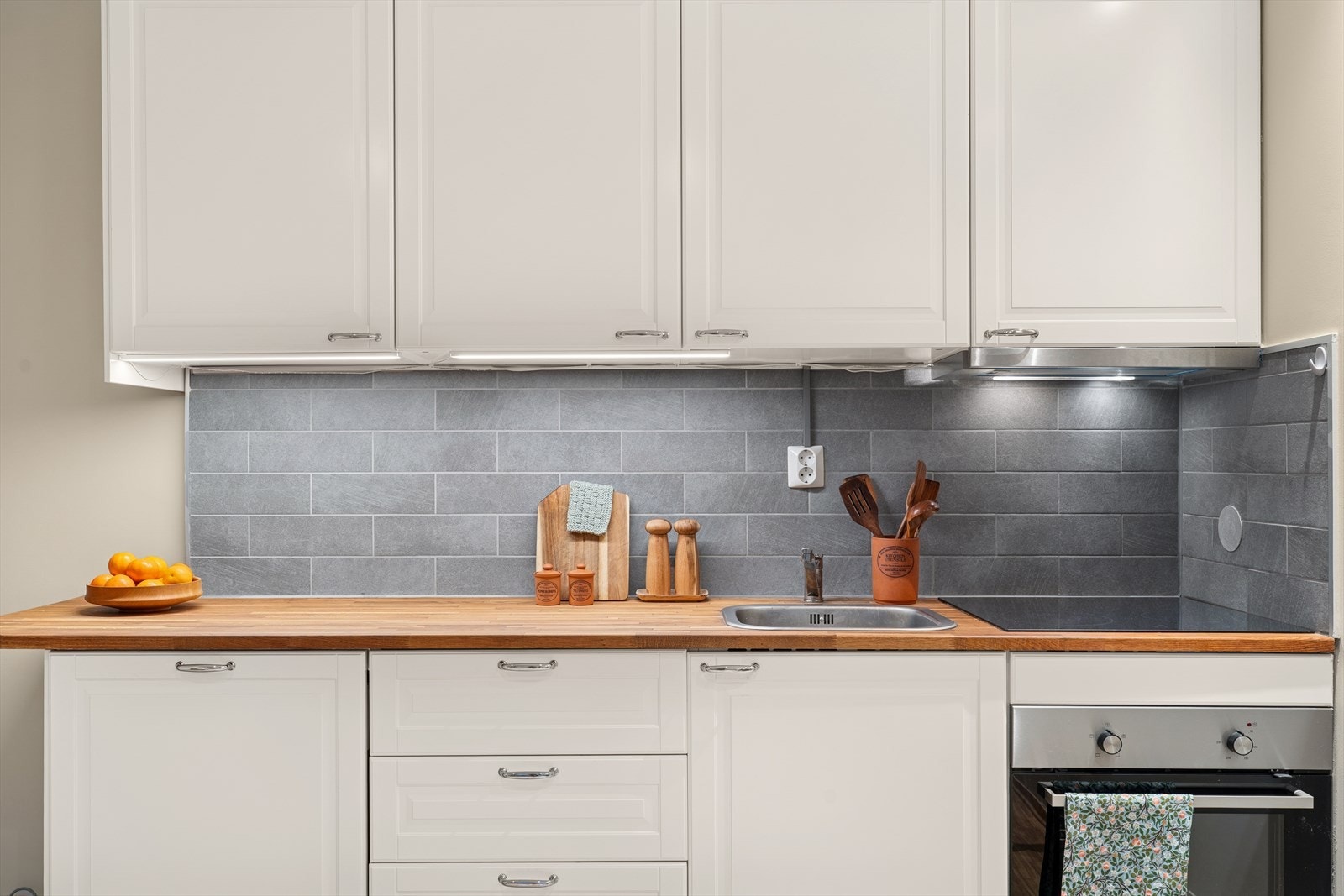 Kjøkkenet er innredet med profilerte fronter, benkeplate med nedfelt kum, fliser over benken og belysning under overskapene Galleribilde