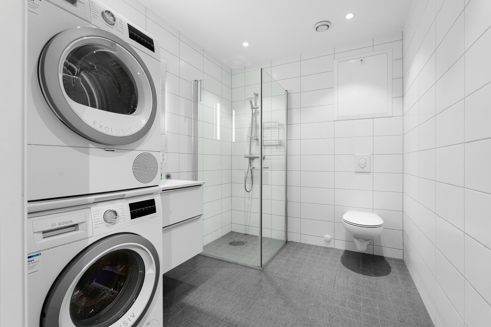 Pent bad med flislagte overflater og gulvvarme, dusjhjørne, veggmonter toalett med skjult sisterne og servant i skap. Galleribilde